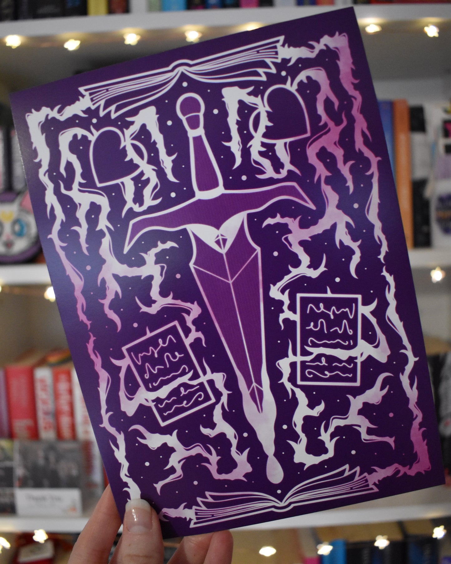 Hand holding print in centre of image at an angle against a blurry bookcase with fairy lights. Purple illustrated design featuring white and pink, with a dagger in the centre, whispy/thunder/thorn branch elements mixed with open books, pages and hearts.