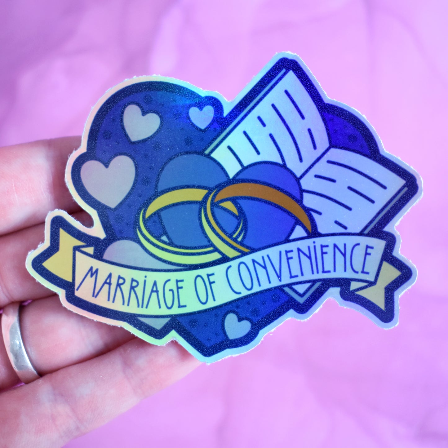 White hand holding sticker. Sticker design is a big blue heart with smaller hearts, an open book and a pair of gold wedding rings inside it. A pale yellow banner across the middle of the design reads marriage of convenience. Sticker has a border all around the design. Background is blurry/distance of a pinkish watercolour style.
