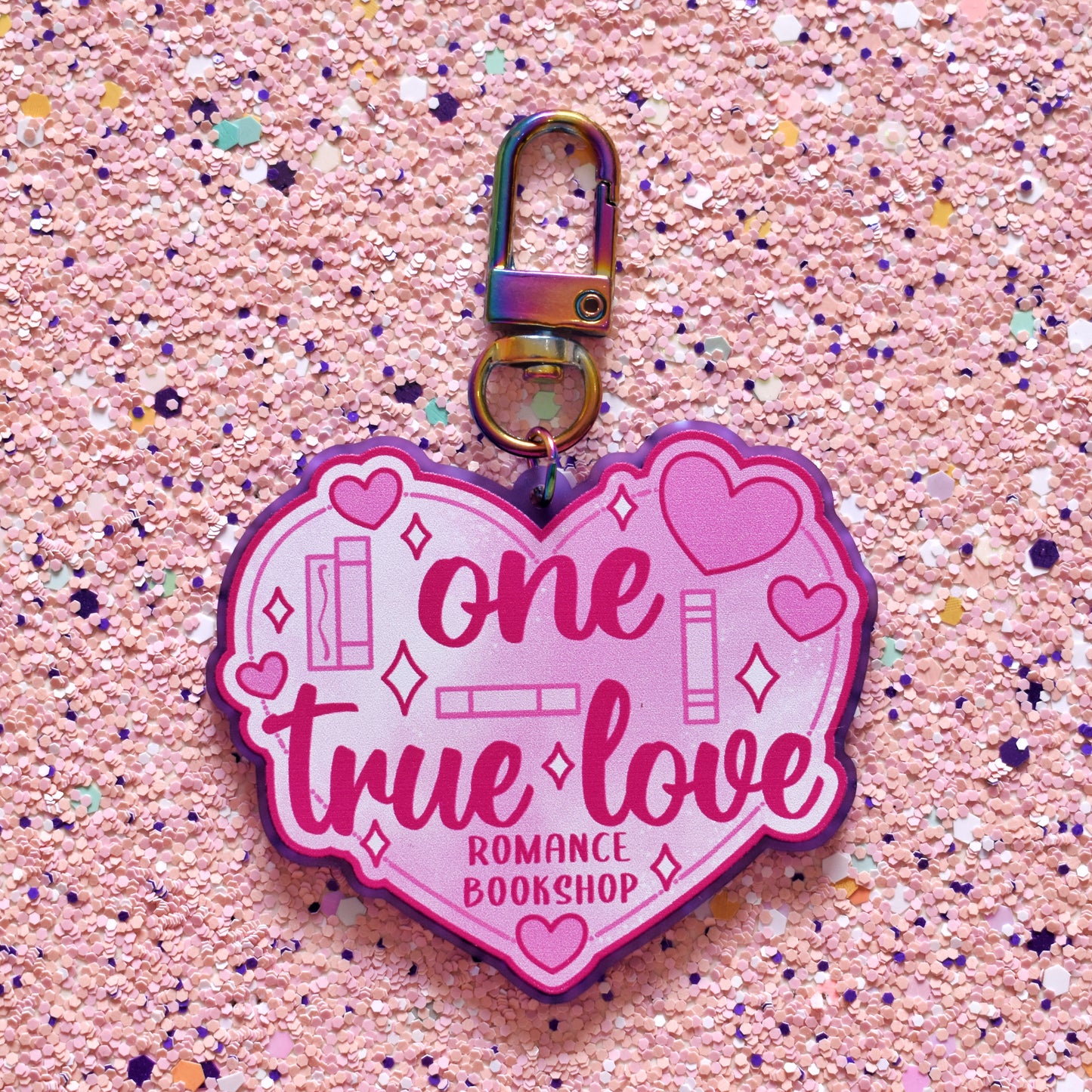 Close up of acrylic keyring in the centre of the image. Featuring a big pink heart with smaller hearts and book spines across the design. Design reads one true love romance bookshop. Keyring is printed on frosted purple acrylic with a rainbow clasp. The background of the image is peach soft sparkly.