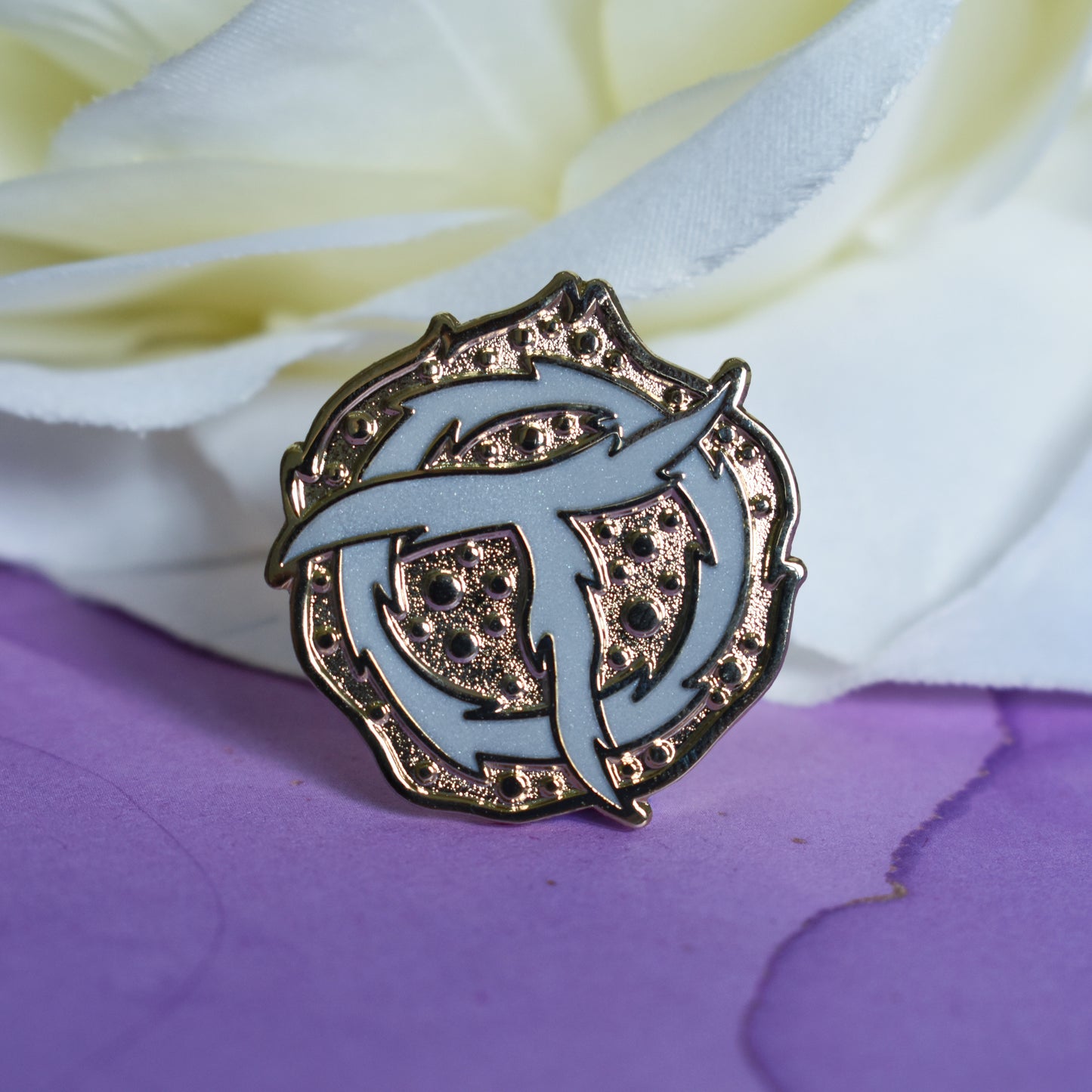 Gold metal enamel pin in centre of image. Enamel pin is roughly round with raised and textured recessed metal. In the centre is a rough pearl white ring with a rough pearl white T shape on top. Background of photograph is purple watercolour style with a fake cream flower.