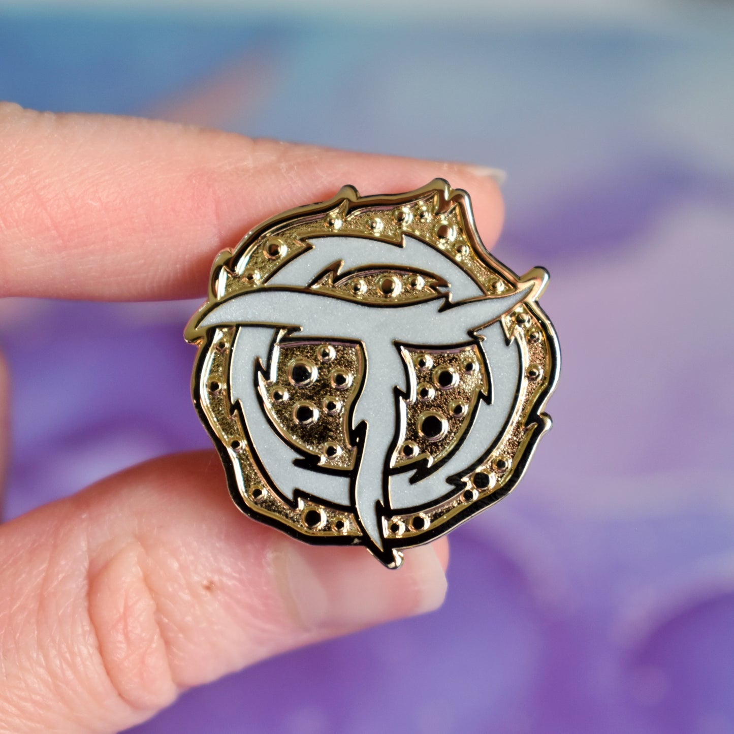 White hand holding gold metal enamel pin. Enamel pin is roughly round with raised and textured recessed metal. In the centre is a rough pearl white ring with a rough pearl white T shape on top. Background of photograph is blue/purple watercolour style.