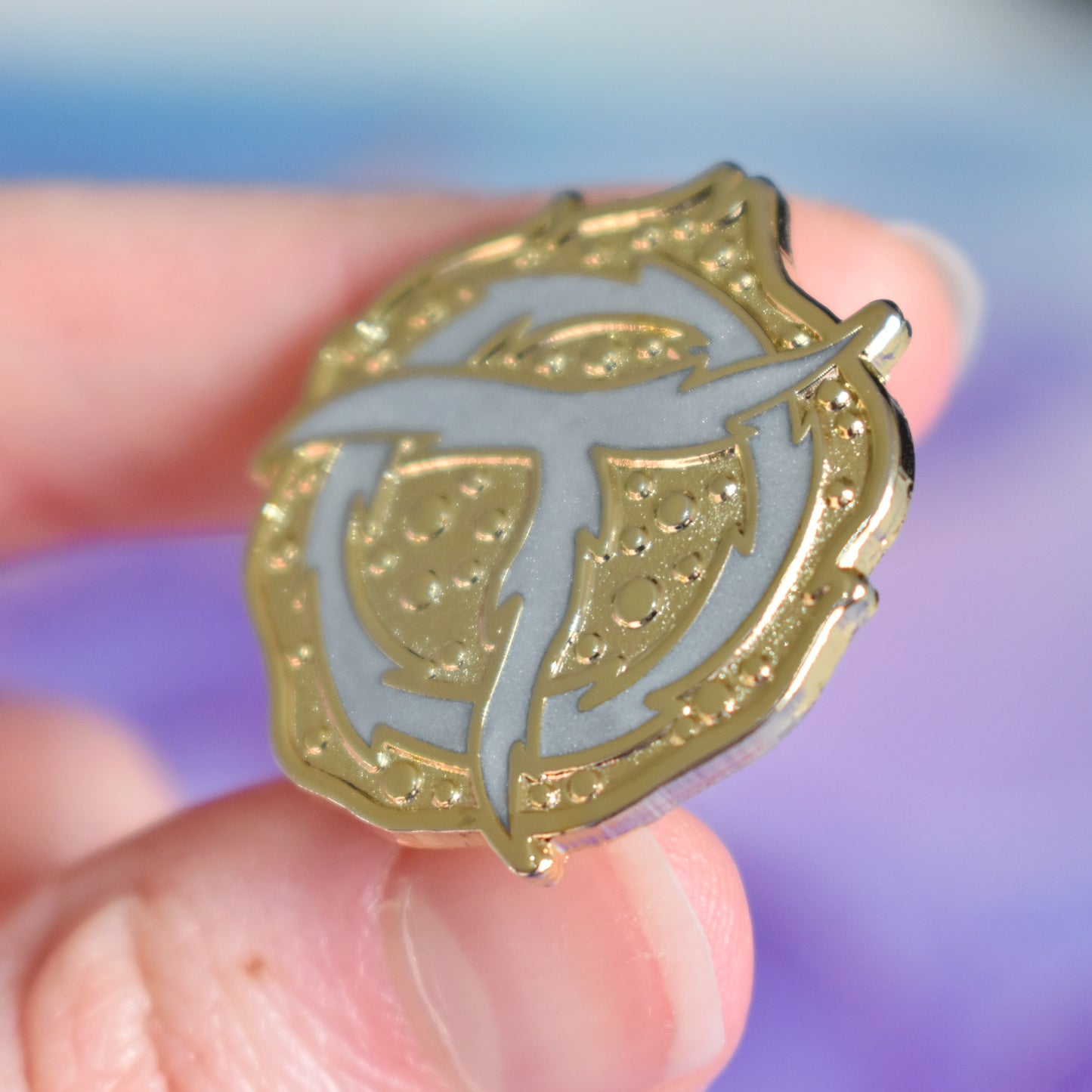 White hand holding gold metal enamel pin tilted to make the metal catch more light. Enamel pin is roughly round with raised and textured recessed metal. In the centre is a rough pearl white ring with a rough pearl white T shape on top. Background of photograph is blue/purple watercolour style.