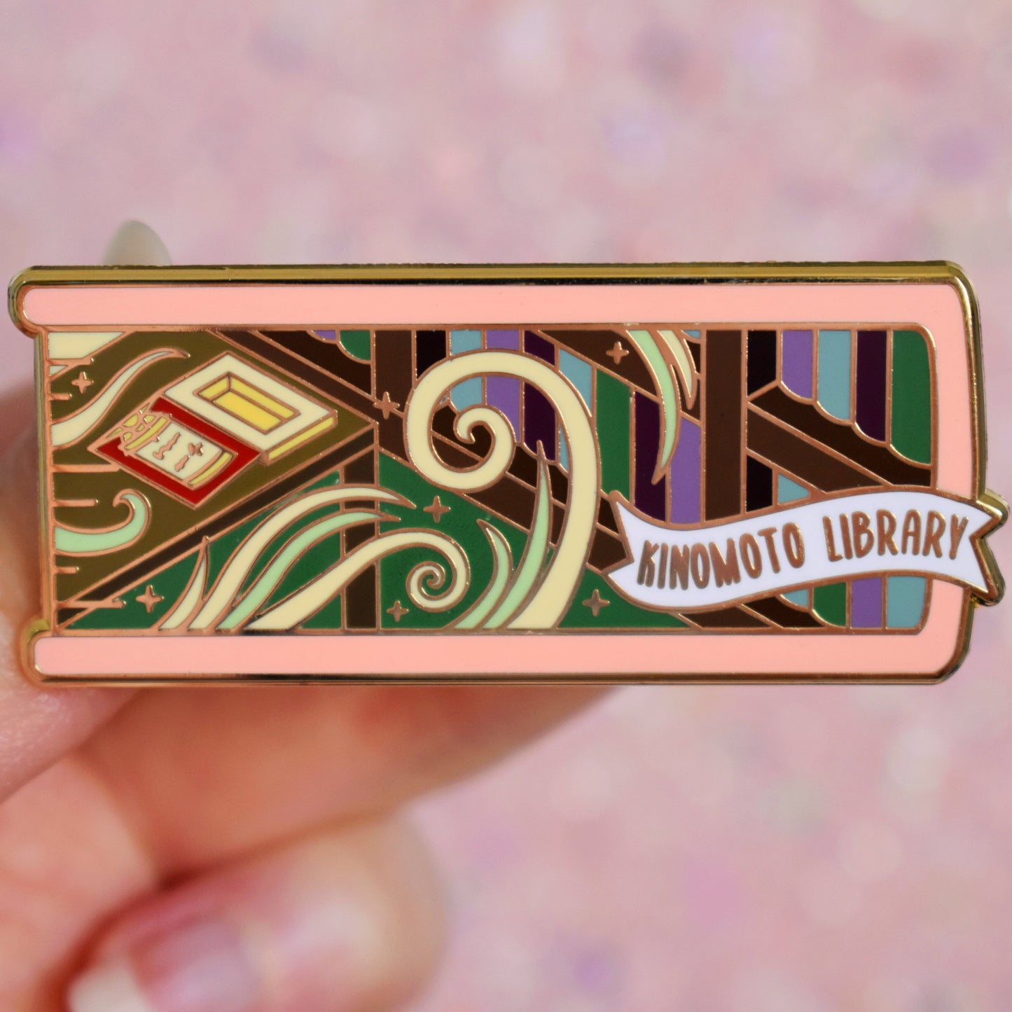 White hand holding gold metal enamel pin. Enamel pin is shaped like the end of a book in light pink and features a scene in a library. Shelves of books on the right with a desk on the left. Desk has an open clow book and swirls of white and green with sparkles. A small banner reads kinomoto library on the bottom right of the design. Background of photograph is peachy coloured.