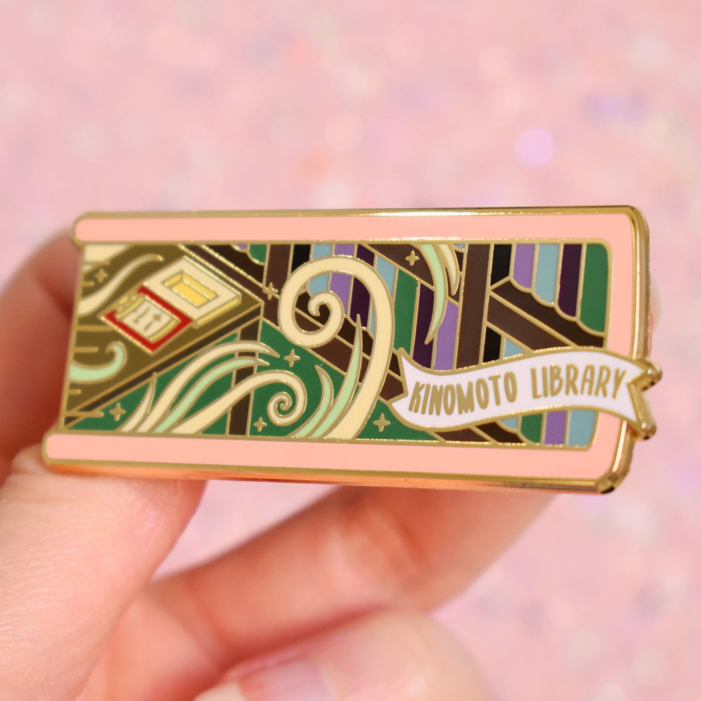White hand holding gold metal enamel pin tilted to make the metal catch more light. Enamel pin is shaped like the end of a book in light pink and features a scene in a library. Shelves of books on the right with a desk on the left. Desk has an open clow book and swirls of white and green with sparkles. A small banner reads kinomoto library on the bottom right of the design. Background of photograph is peachy coloured.
