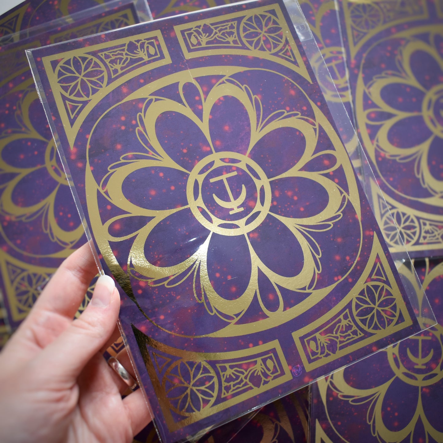 White hand holding print. A5 print featuring a gold foil design with swirls and flower repeating motifs. Background of print is a redish purple cloudy and sparkles design. Background of photograph is the same prints scattered.