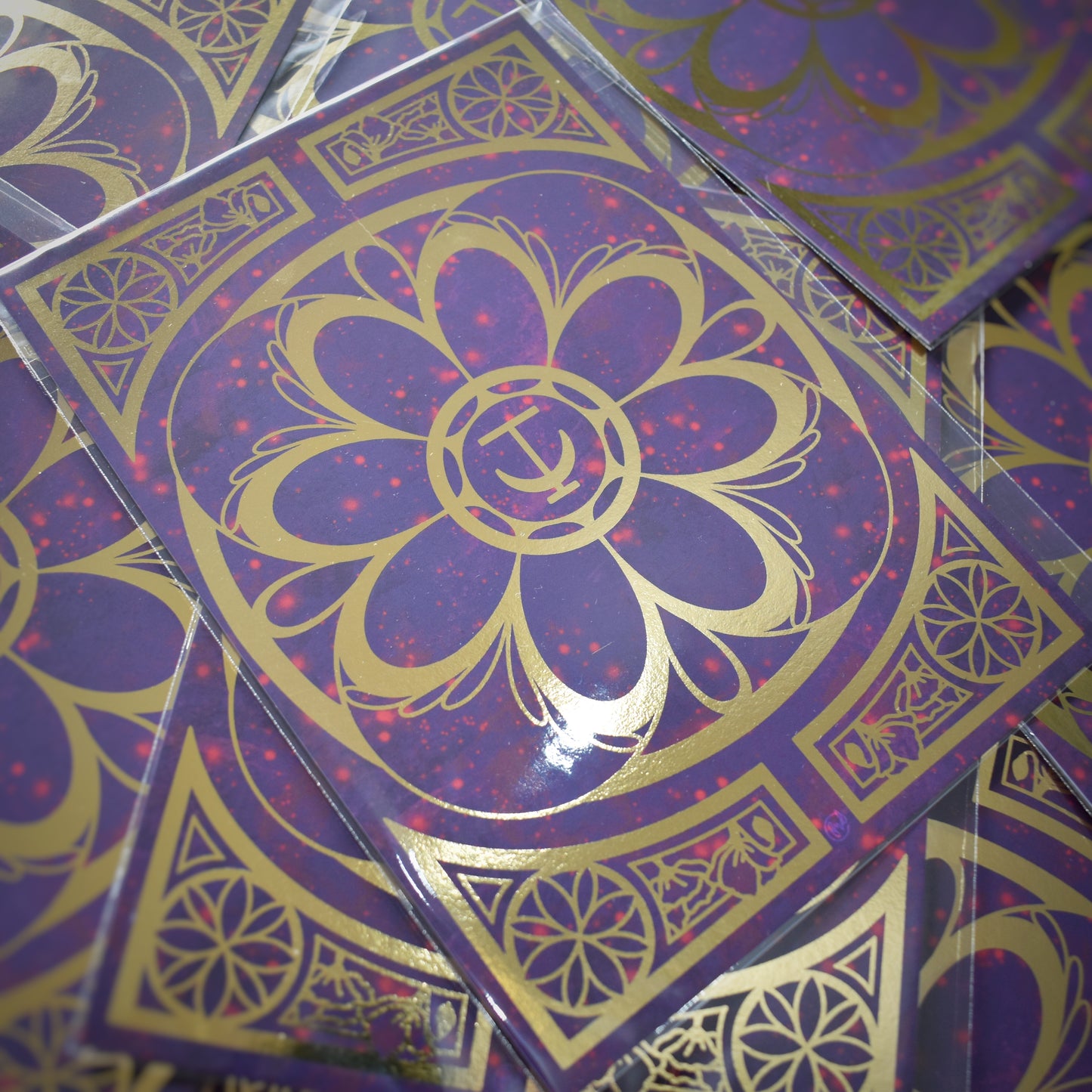 Multiple of same print scattered. A5 print featuring a gold foil design with swirls and flower repeating motifs. Background of print is a redish purple cloudy and sparkles design.