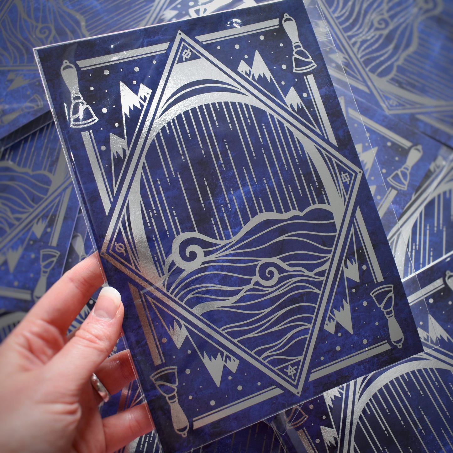 White hand holding print. A5 print featuring a silver foil design with a diamond, water, mountains, bells, some of which are repeating motifs. Background of print is a black and blue cloudy design. Background of photograph is the same prints scattered.