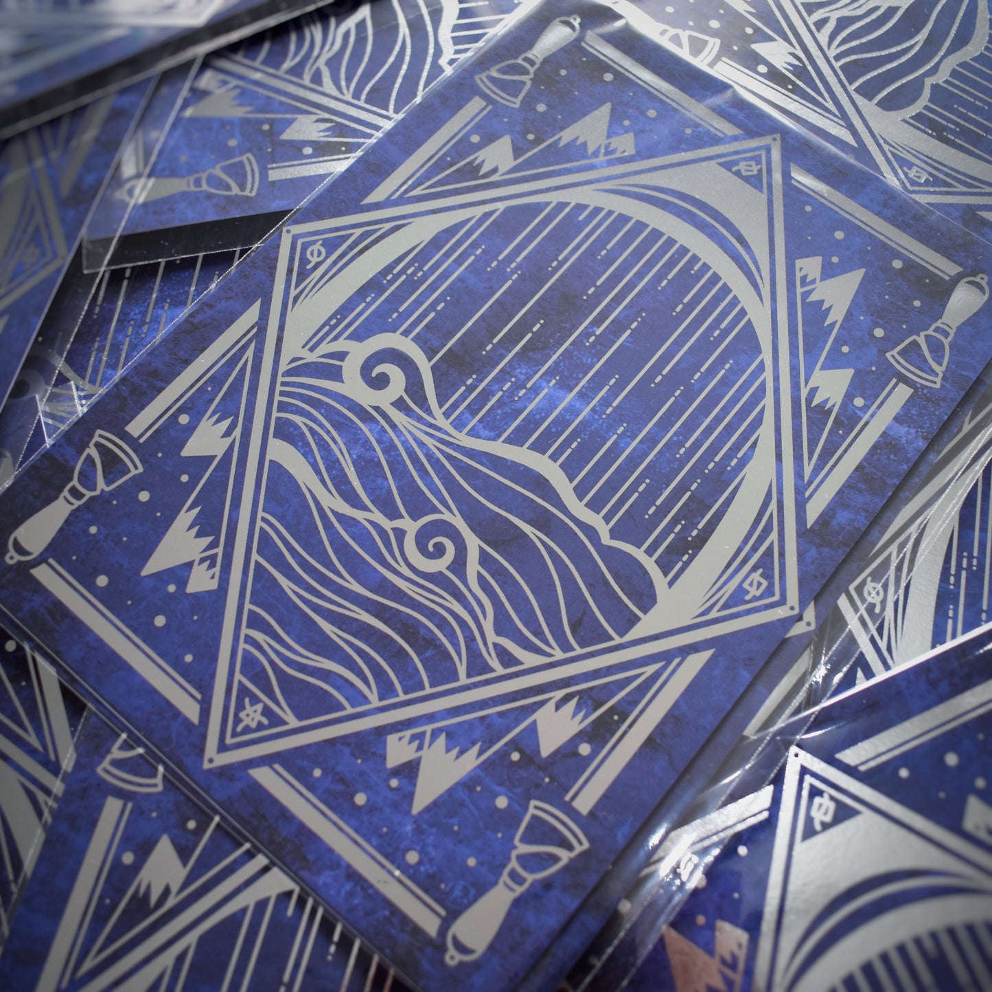 Multiple of same print scattered. A5 print featuring a silver foil design with a diamond, water, mountains, bells, some of which are repeating motifs. Background of print is a black and blue cloudy design.