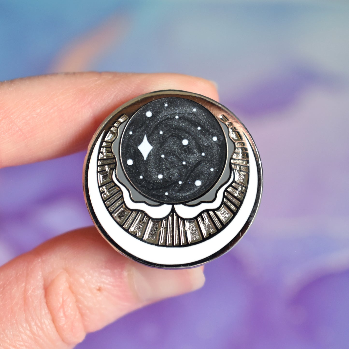 White hand holding silver metal enamel pin. Enamel pin has a swirly black pearl circle top centre with some sparkles on. under is a cresent moon shape with raised metal liners and textured recessed metal areas. Under that and up the sides is a white cresent moon. Background of photograph is blurry purple/blue coloured.