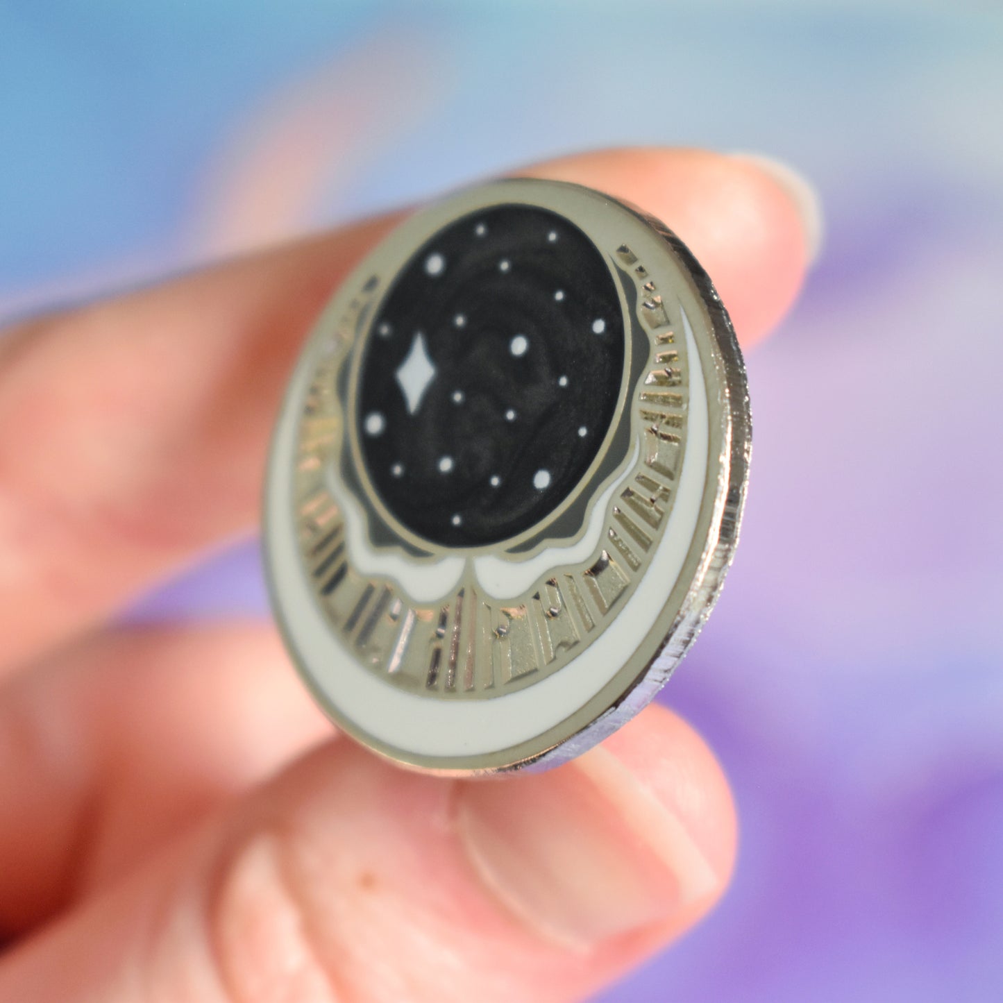 White hand holding silver metal enamel pin tilted to make the metal catch more light. Enamel pin has a swirly black pearl circle top centre with some sparkles on. under is a cresent moon shape with raised metal liners and textured recessed metal areas. Under that and up the sides is a white cresent moon. Background of photograph is blurry purple/blue coloured.