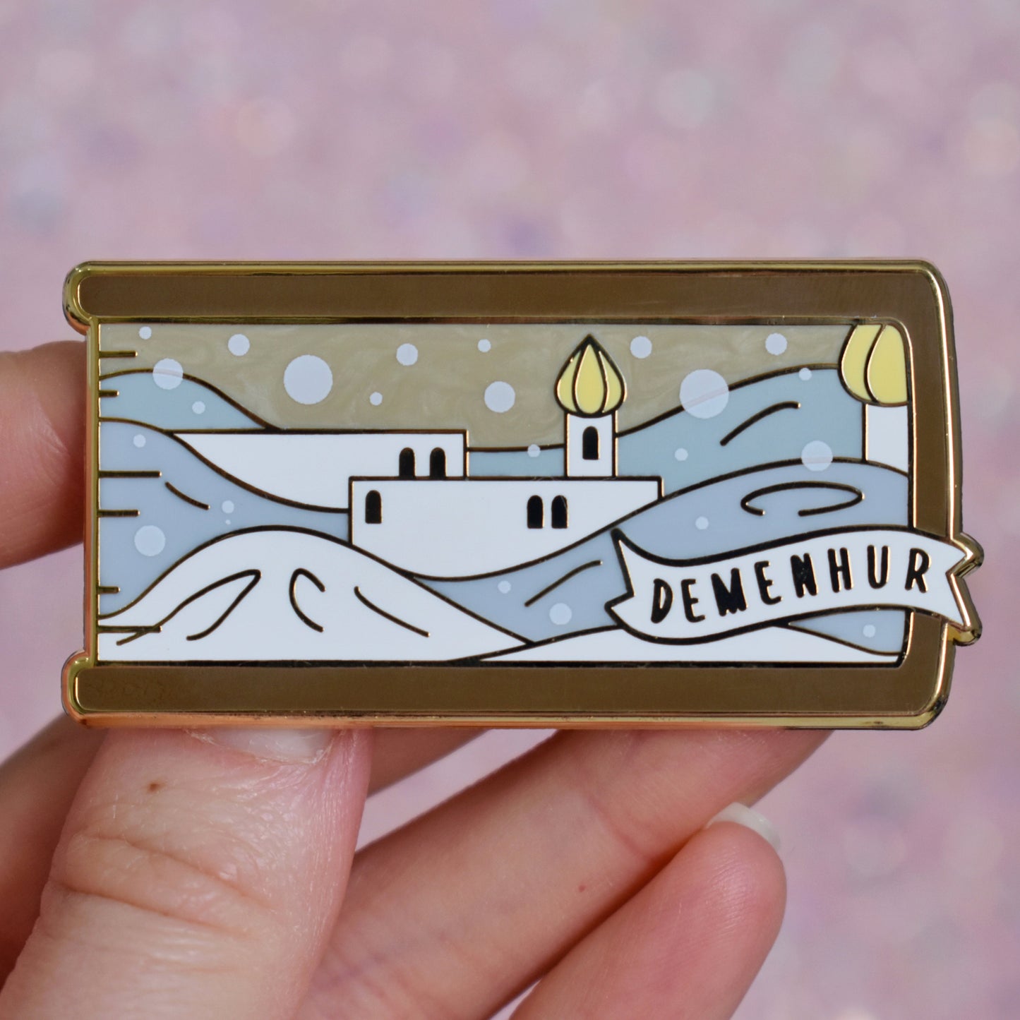 White hand holding gold metal enamel pin. Enamel pin is shaped like the end of a book in brown and features a white adn grey pearl snowy scene with white buildings and yellow details. The sky is a sandy pearl. A small banner reads demenhur on the bottom right of the design. Background of photograph is grey pinkish coloured.