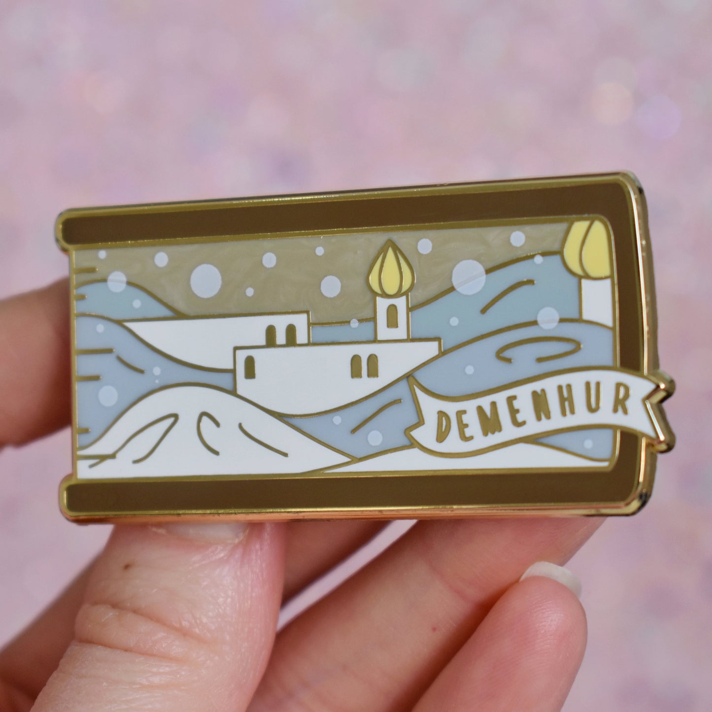 White hand holding gold metal enamel pin tilted to make the metal catch more light. Enamel pin is shaped like the end of a book in brown and features a white adn grey pearl snowy scene with white buildings and yellow details. The sky is a sandy pearl. A small banner reads demenhur on the bottom right of the design. Background of photograph is grey pinkish coloured.