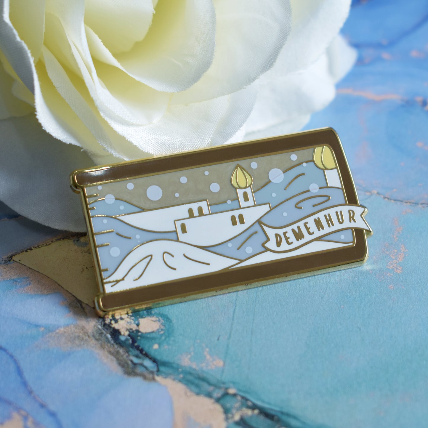 Gold metal enamel pin in centre of image. Enamel pin is shaped like the end of a book in brown and features a white adn grey pearl snowy scene with white buildings and yellow details. The sky is a sandy pearl. A small banner reads demenhur on the bottom right of the design. Background of photograph is blue water inspired with a fake cream flower.