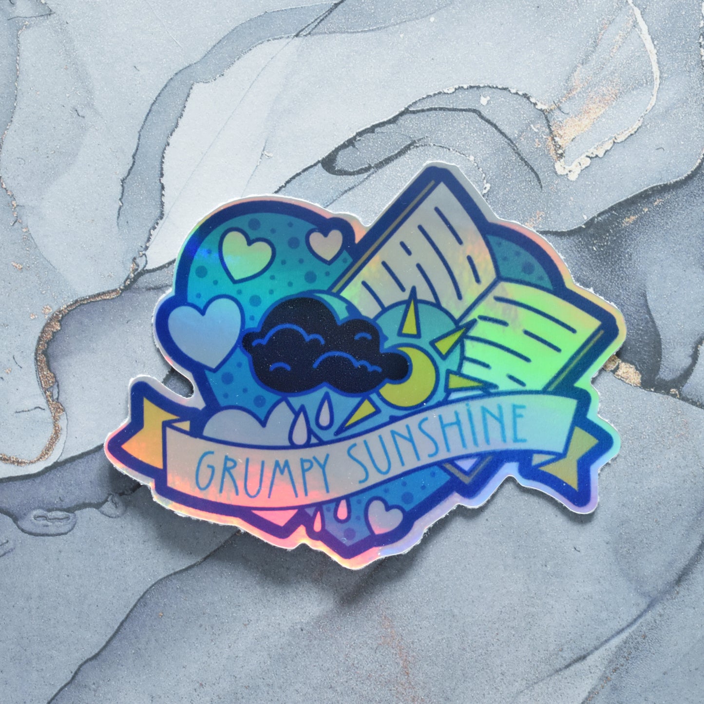 Sticker design is a big blue heart with smaller hearts, an open book and dark cloud with sun peaking out from beside the cloud inside the big heart. A pale yellow banner across the middle of the design reads grumpy sunshine. Sticker has a border all around the design. Holographic effects visible across design. Design has uneven border and blurry print on right side. Background is a grey watercolour style.