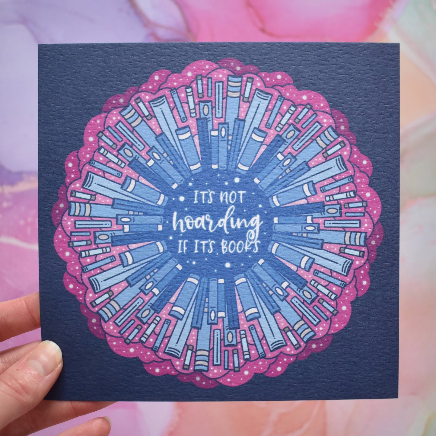 White hand holding print. Square print featuring a repeating pattern of blue bookspines and pink cloud/swirls with darker blue around the edge. Design reads it's not hoarding if it's books in white/light blue at the centre of the design. Background of photograph is pink/lilac/peach watercolour style.