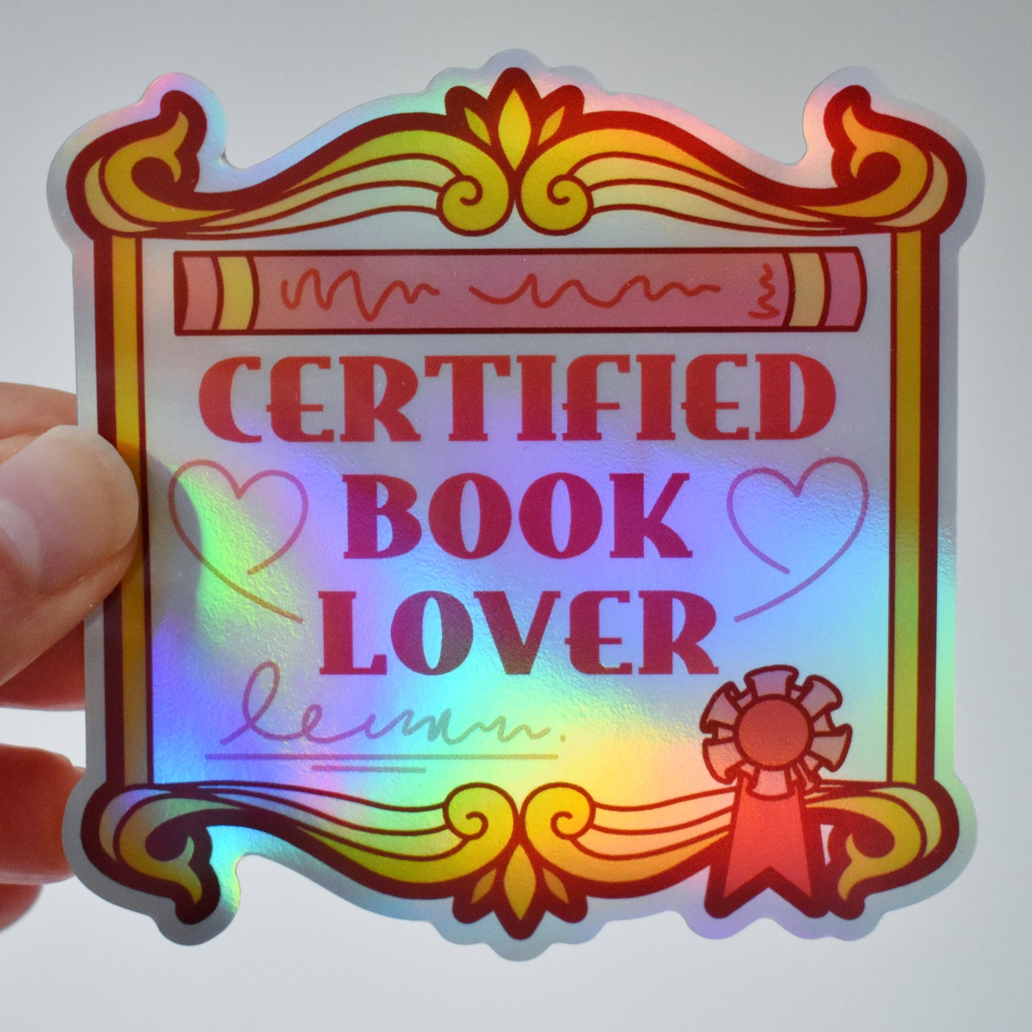 Holographic sticker with 'Certified Book Lover' text held by a hand on an almost white background