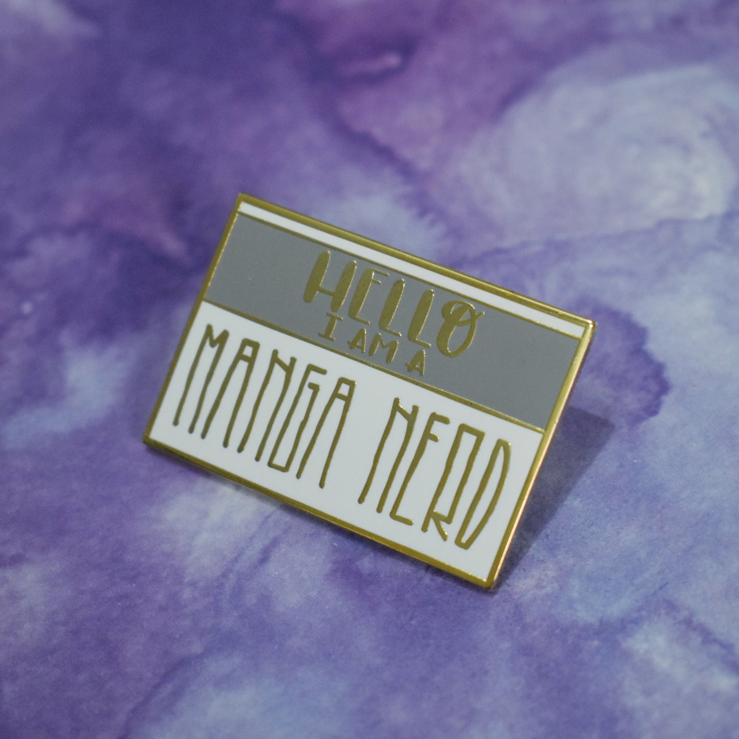 Rectangle gold metal enamel pin at an angle in center of photo. White background with a grey band across most of top half. Hello I am a manga nerd written across design. Manga nerd in a large skinny squared sans serif style font. Background of photo is purple watercolour style.