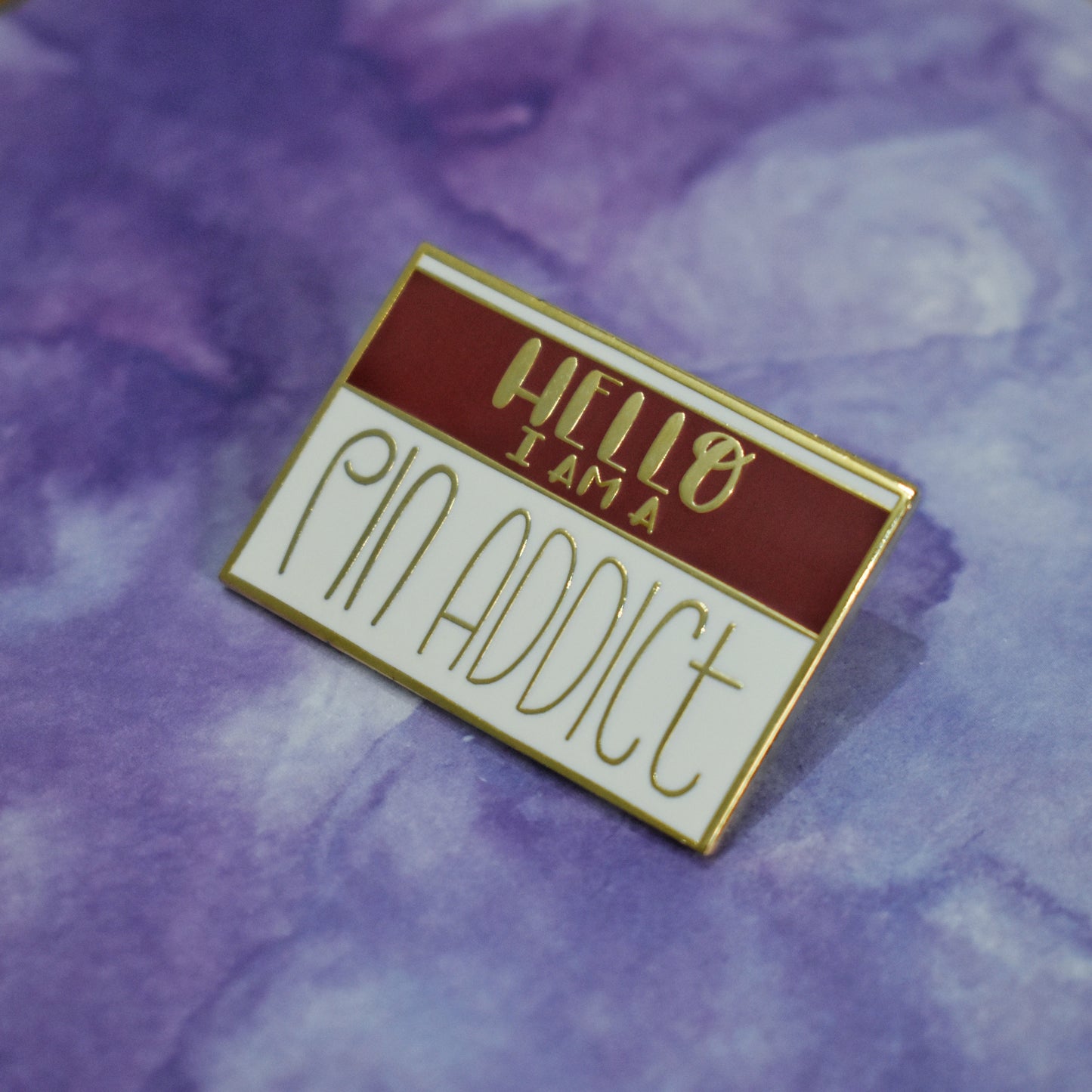 Rectangle gold metal enamel pin at an angle in center of photo. White background with a maroon band across most of top half. Hello I am a pin addict written across design. Pin addict in a large skinny curved sans serif style font. Background of photo is purple watercolour style.