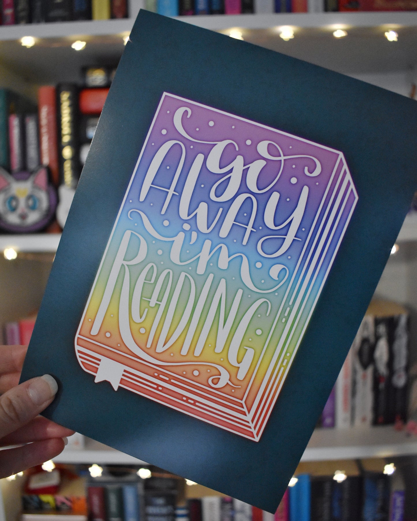 Hand holding print in centre of image at an angle against a blurry bookcase with fairy lights. Teal illustration design with a rainbow book in the centre, facing forwards. Go away I'm reading n handwritten font in white ontop of the rainbow book.
