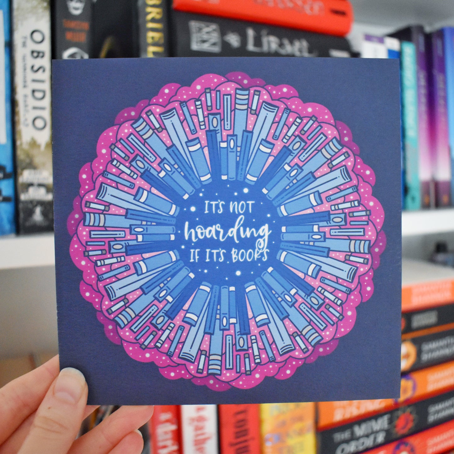 White hand holding print. Square print featuring a repeating pattern of blue bookspines and pink cloud/swirls with darker blue around the edge. Design reads it's not hoarding if it's books in white/light blue at the centre of the design. Background of photograph has blurry bookshelf/books.