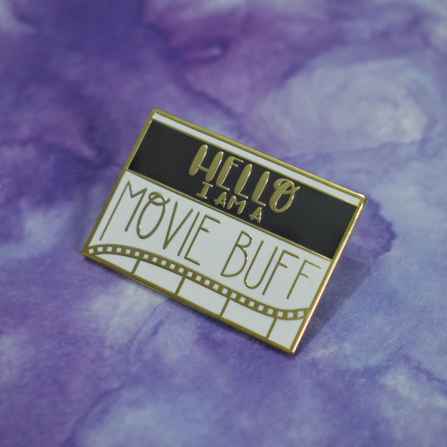 Rectangle gold metal enamel pin at an angle in center of photo. White background with a black band across most of top half. Hello I am a movie buff written across design with a wavy line of old film tape in gold underneath. Movie Buff in a large straight/pointed skinny sans serif style font. Background of photo is purple watercolour style.
