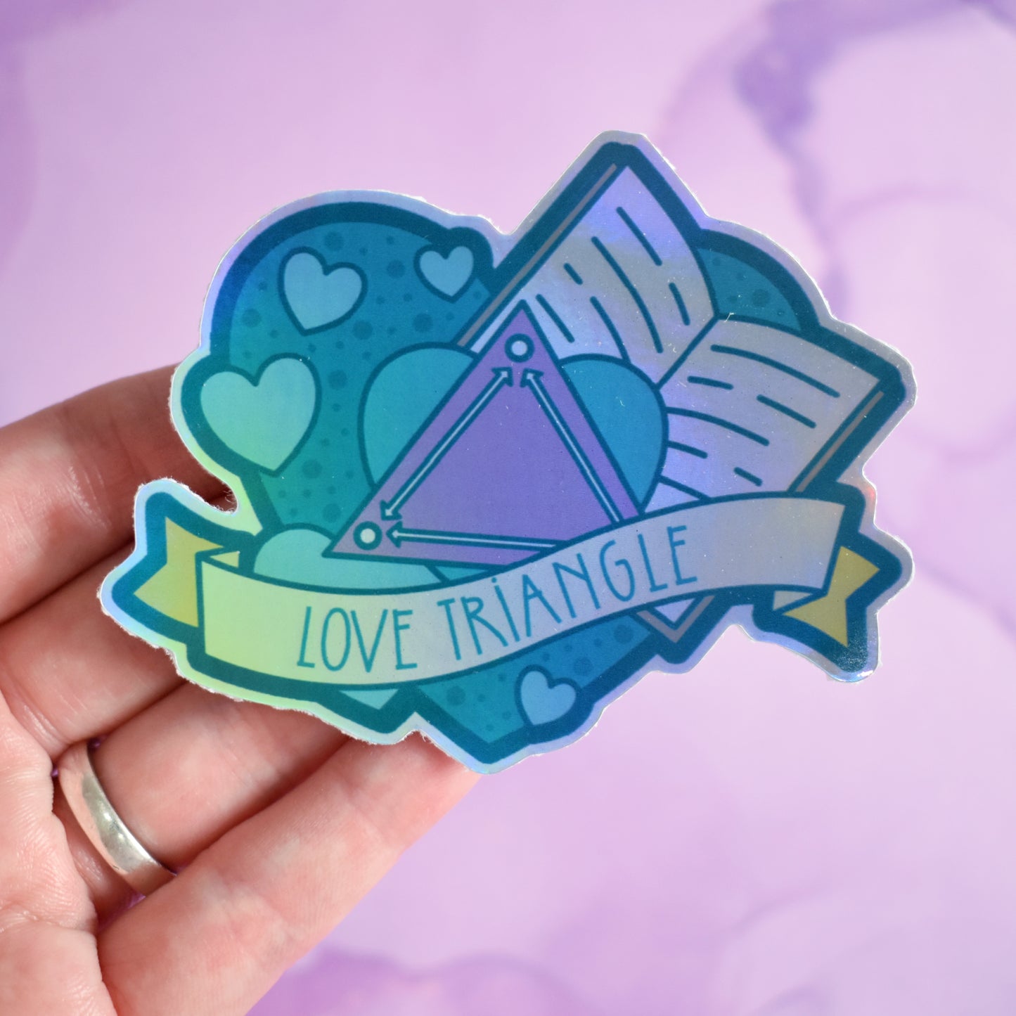 White hand holding sticker. Sticker design is a big green/teal heart with smaller hearts, an open book and a pink triangle with arrows pointing to each corner inside it. A pale yellow banner across the middle of the design reads love triangle. Sticker has a border all around the design. Background is blurry/distance of a pinkish watercolour style.