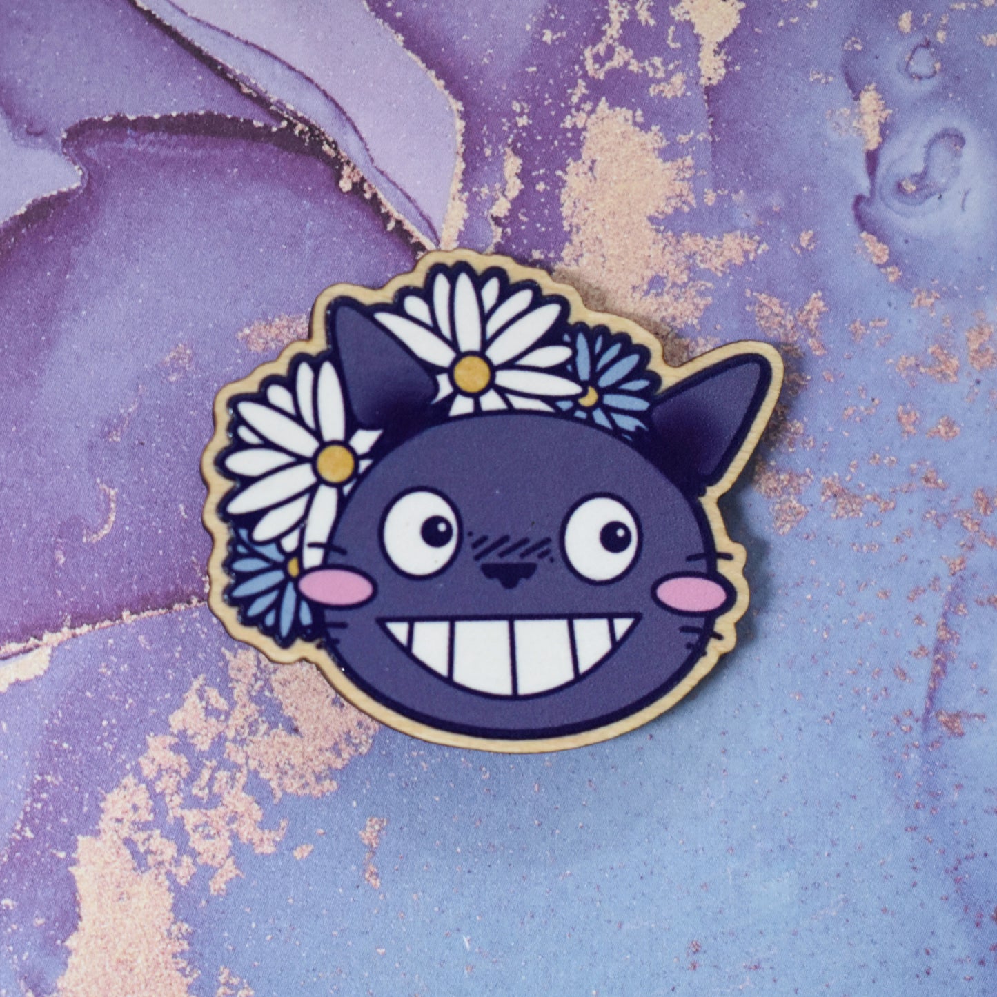 Wooden pin in centre of photo. Design is a close up of the character Totoro's face, with a big smile and cheek blushes. 2 white & 2 blue daisy like flowers are partially behind to the top left of the design. Wooden pin has a gloss so has a little shiny around the edge. Background is soft watercolour blue and purple style.