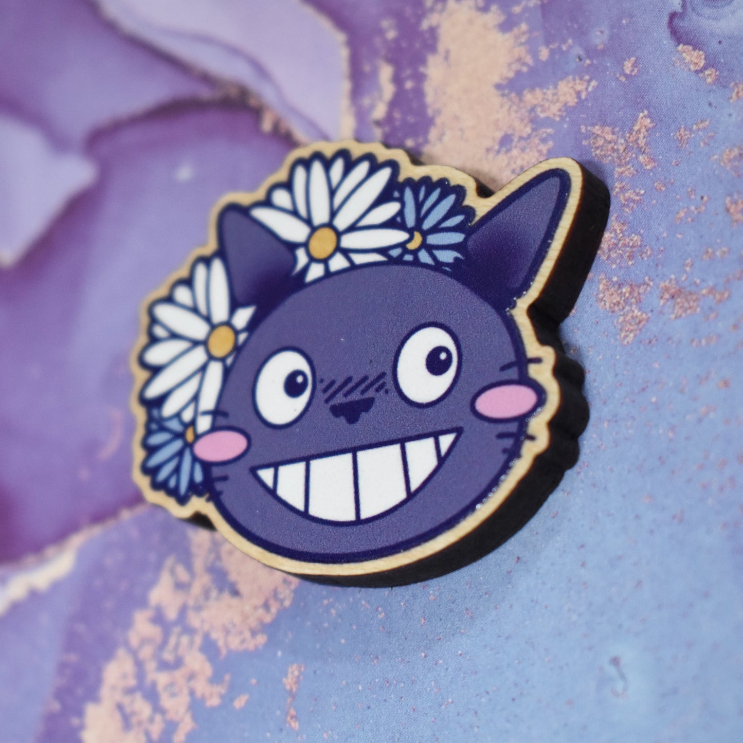 Wooden pin in centre of photo at an angle, which shows the cut edge of the pin which is darker than the wood visable on the front. Design is a close up of the character Totoro's face, with a big smile and cheek blushes. 2 white & 2 blue daisy like flowers are partially behind to the top left of the design. Wooden pin has a gloss so has a little shiny around the edge. Background is soft watercolour blue and purple style.