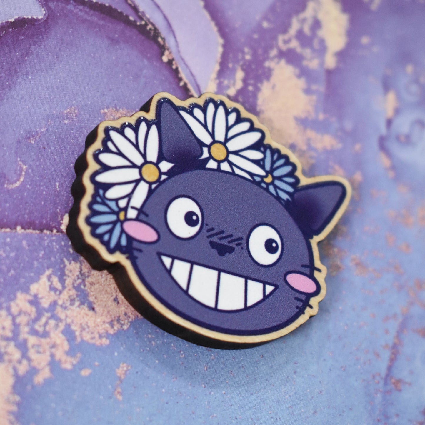 Wooden pin in centre of photo at an angle, which shows the cut edge of the pin which is darker than the wood visable on the front. Design is a close up of the character Totoro's face, with a big smile and cheek blushes. 2 white & 2 blue daisy like flowers are partially behind to the top left of the design. Wooden pin has a gloss so has a little shiny around the edge. Background is soft watercolour blue and purple style.