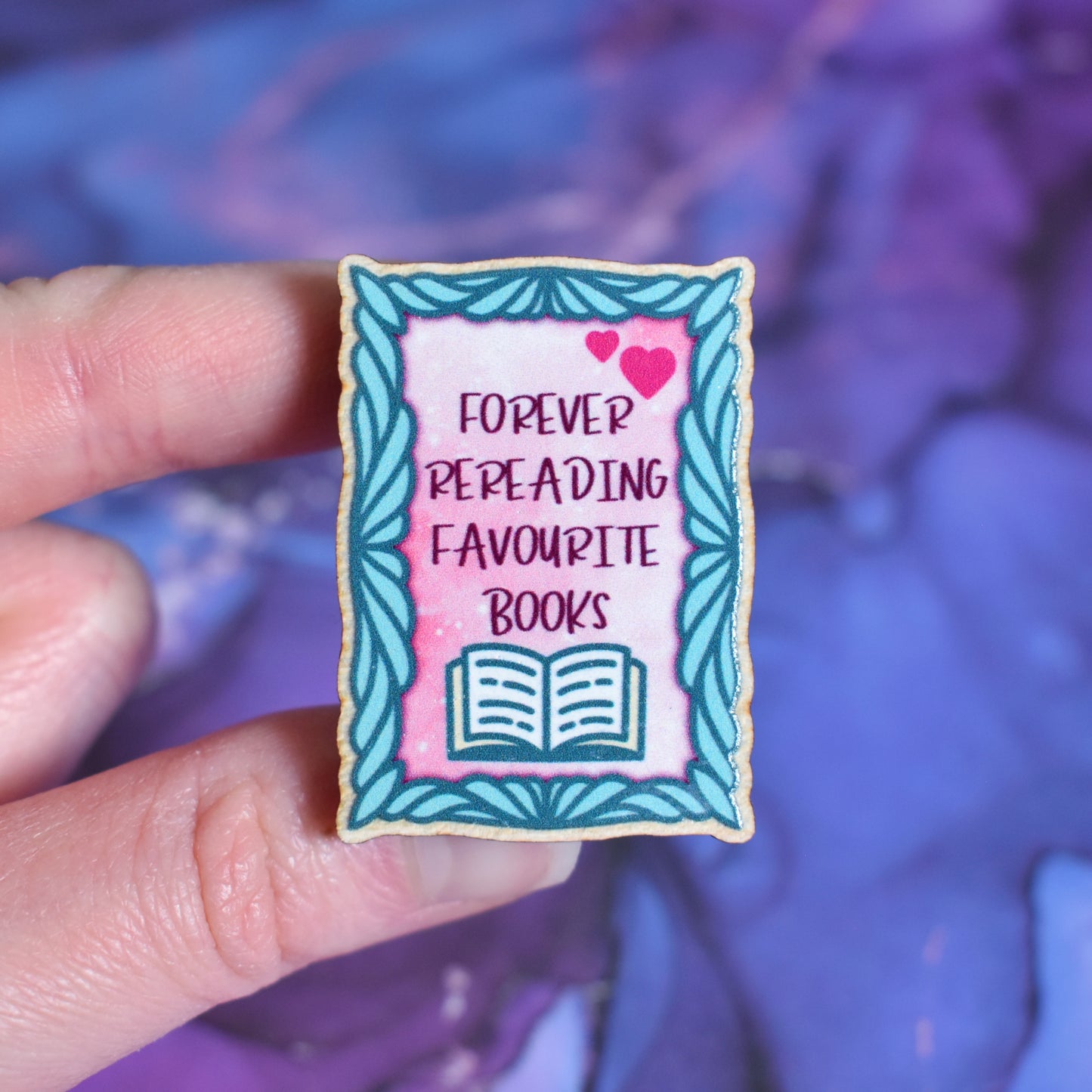 White hand holding small rectangular wooden pin in centre of photo. Design features a curved teal frame with a pink centre. In the centre is 2 pink hearts and written forever rereading favourite books with and open book underneath. Wooden pin has a gloss so has a little shiny around the edge. Background is soft watercolour blue and purple style.