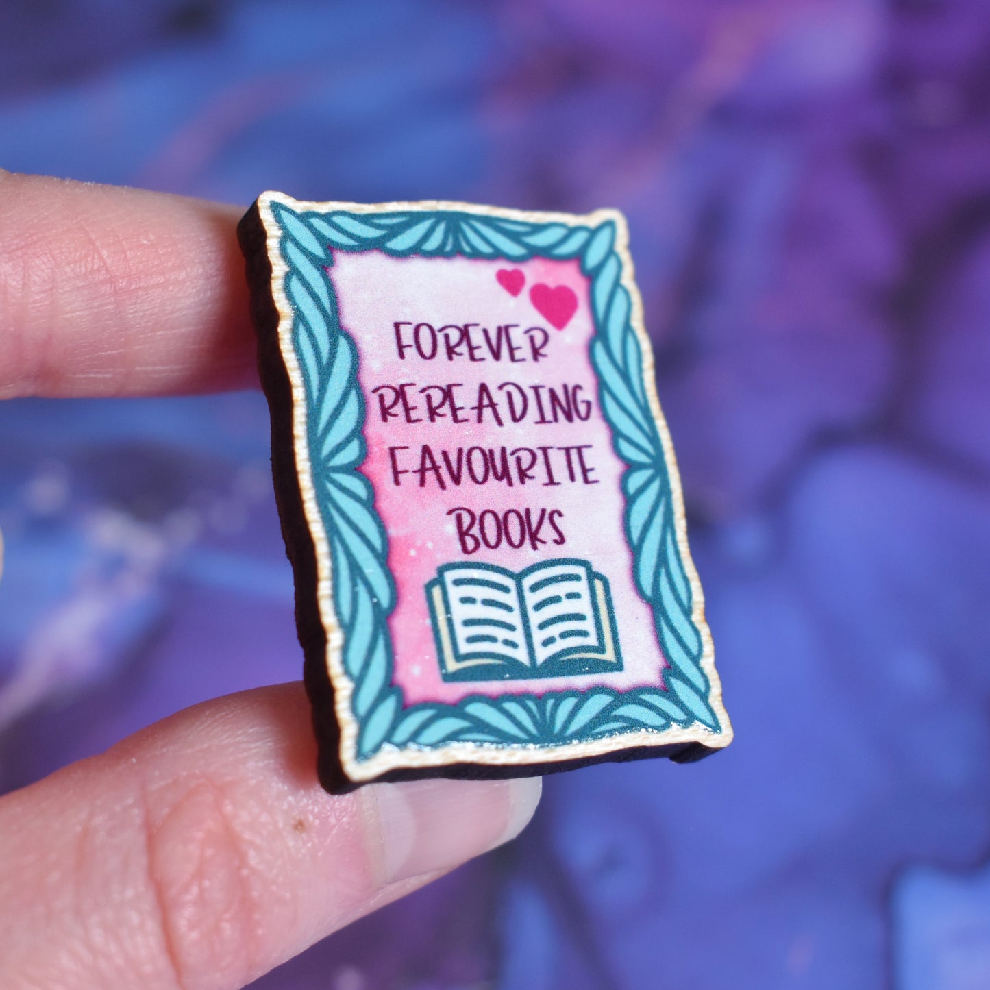 White hand holding small rectangular wooden pin in centre of photo at an angle, which shows the cut edge of the pin which is darker than the wood visable on the front. Design features a curved teal frame with a pink centre. In the centre is 2 pink hearts and written forever rereading favourite books with and open book underneath. Wooden pin has a gloss so has a little shiny around the edge. Background is soft watercolour blue and purple style.