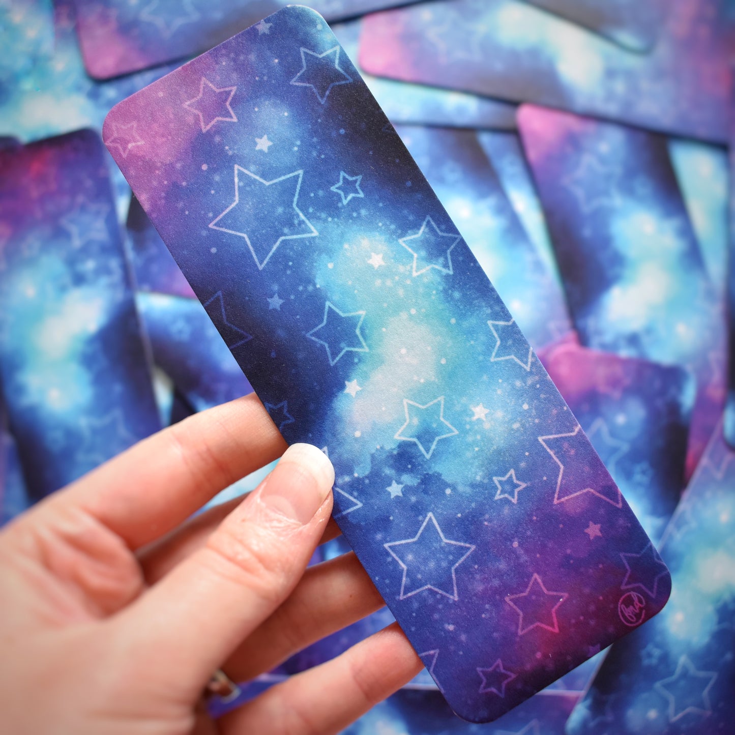 White hand holding bookmark. Bookmark with rounded edges is a galaxy blend of pink, purple, blue and teal, with pale hollow and filled in stars. Bookmark has a slight sparkle/sheen. Background is blurry/distance of same stickers.