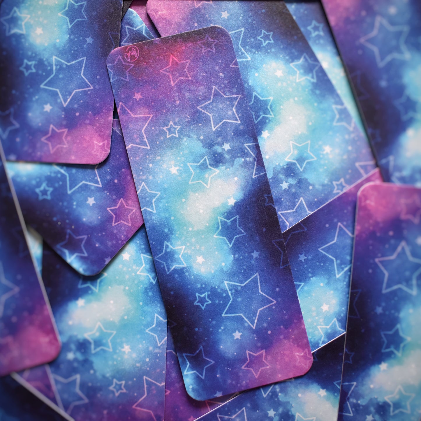 Multiple of same bookmark scattered. Bookmark with rounded edges is a galaxy blend of pink, purple, blue and teal, with pale hollow and filled in stars. Bookmark has a slight sparkle/sheen.