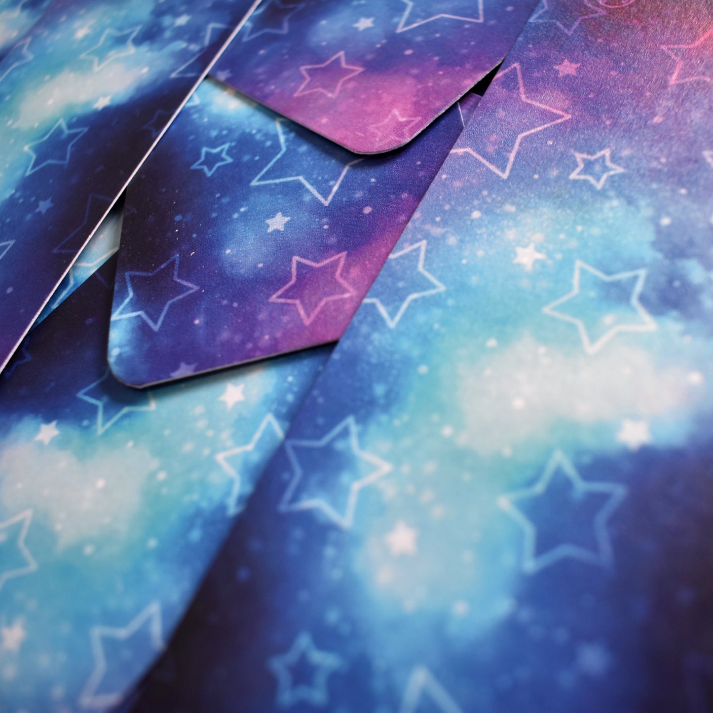 Close up of multiple of same bookmark scattered. Bookmark with rounded edges is a galaxy blend of pink, purple, blue and teal, with pale hollow and filled in stars. Bookmark has a slight sparkle/sheen.
