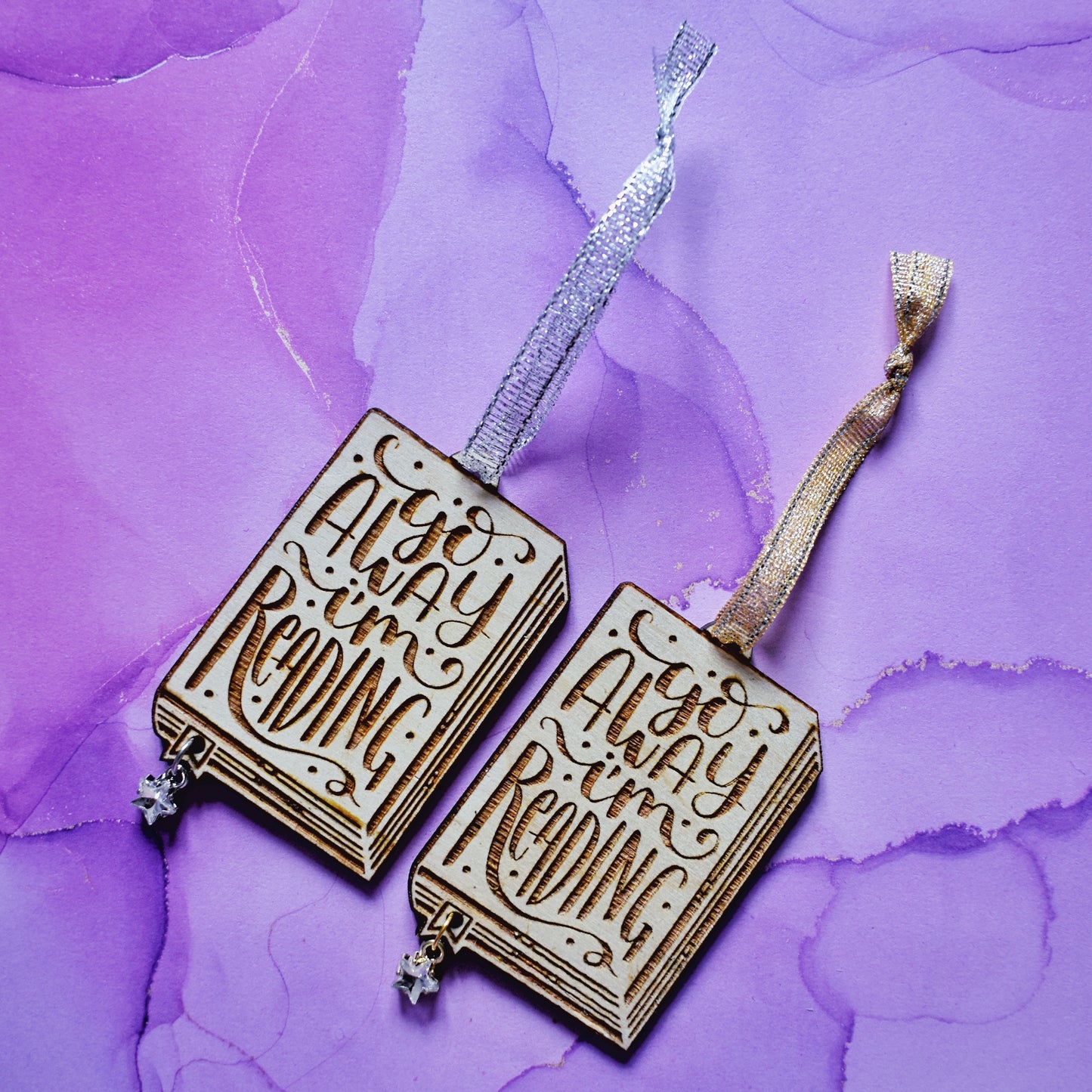 2 wooden ornaments next to each other, at an angle on a purple watercolour background. Ornaments are in the shape of a book with go away I'm reading in loopy writing, showing the page edges at the side and bottom, with a small bookmark end sticking out. A hole in the bookmark end allows for the attachment of a small clear star gem. Left ornamant has a silver ribbon and silver loop on star gem. Right ornamant has a gold ribbon and gold loop on star gem.