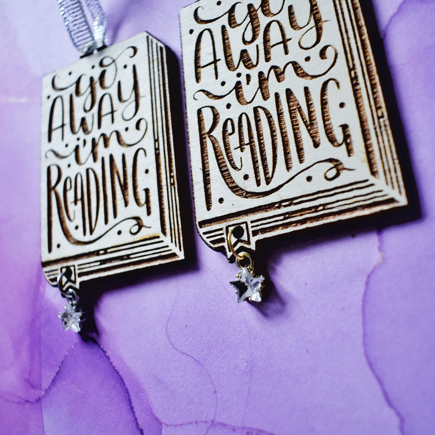 Close up of 2 wooden ornaments next to each other, at an angle on a purple watercolour background. Ornaments are in the shape of a book with go away I'm reading in loopy writing, showing the page edges at the side and bottom, with a small bookmark end sticking out. A hole in the bookmark end allows for the attachment of a small clear star gem. Left ornamant has a silver ribbon and silver loop on star gem. Right ornamant has a gold ribbon and gold loop on star gem.