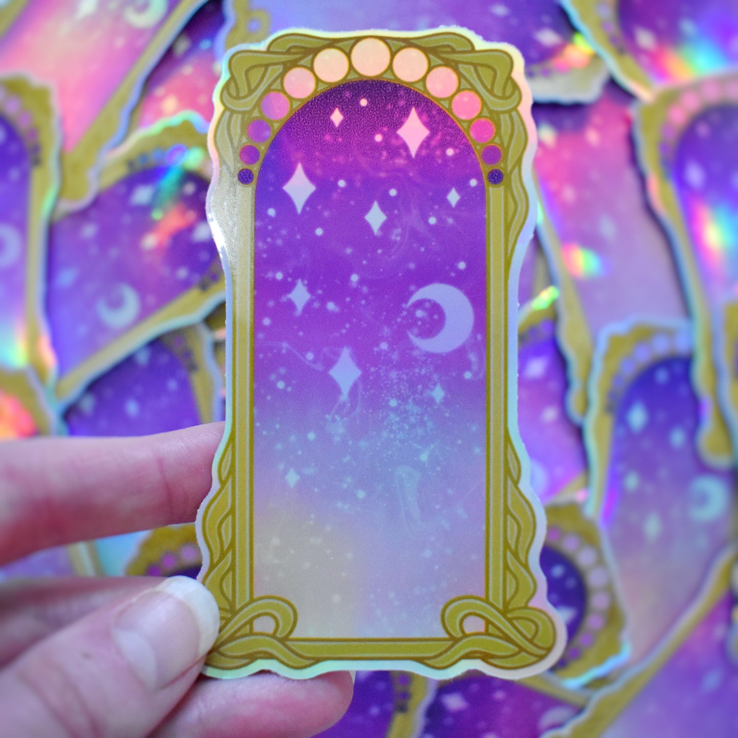 White hand holding sticker. Sticker design is a gold/yellow curly frame with a gradient of circles across the top, from purple to pink. Centre of the design is a from purple to pink gradient with white/hazy stars and moon. Background is blurry/distance of same stickers.