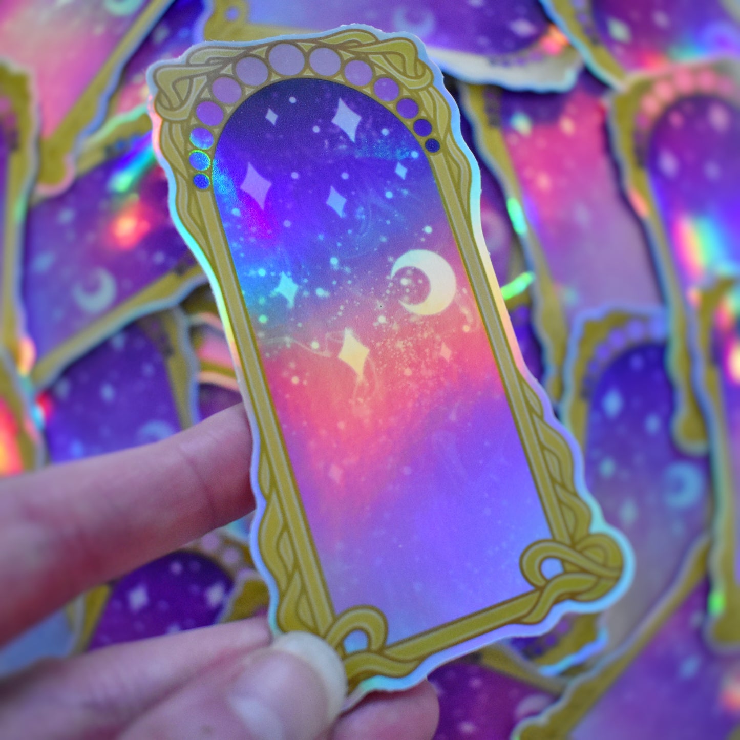 White hand holding sticker tilted to show more holographic effect. Sticker design is a gold/yellow curly frame with a gradient of circles across the top, from purple to pink. Centre of the design is a from purple to pink gradient with white/hazy stars and moon. Background is blurry/distance of same stickers.