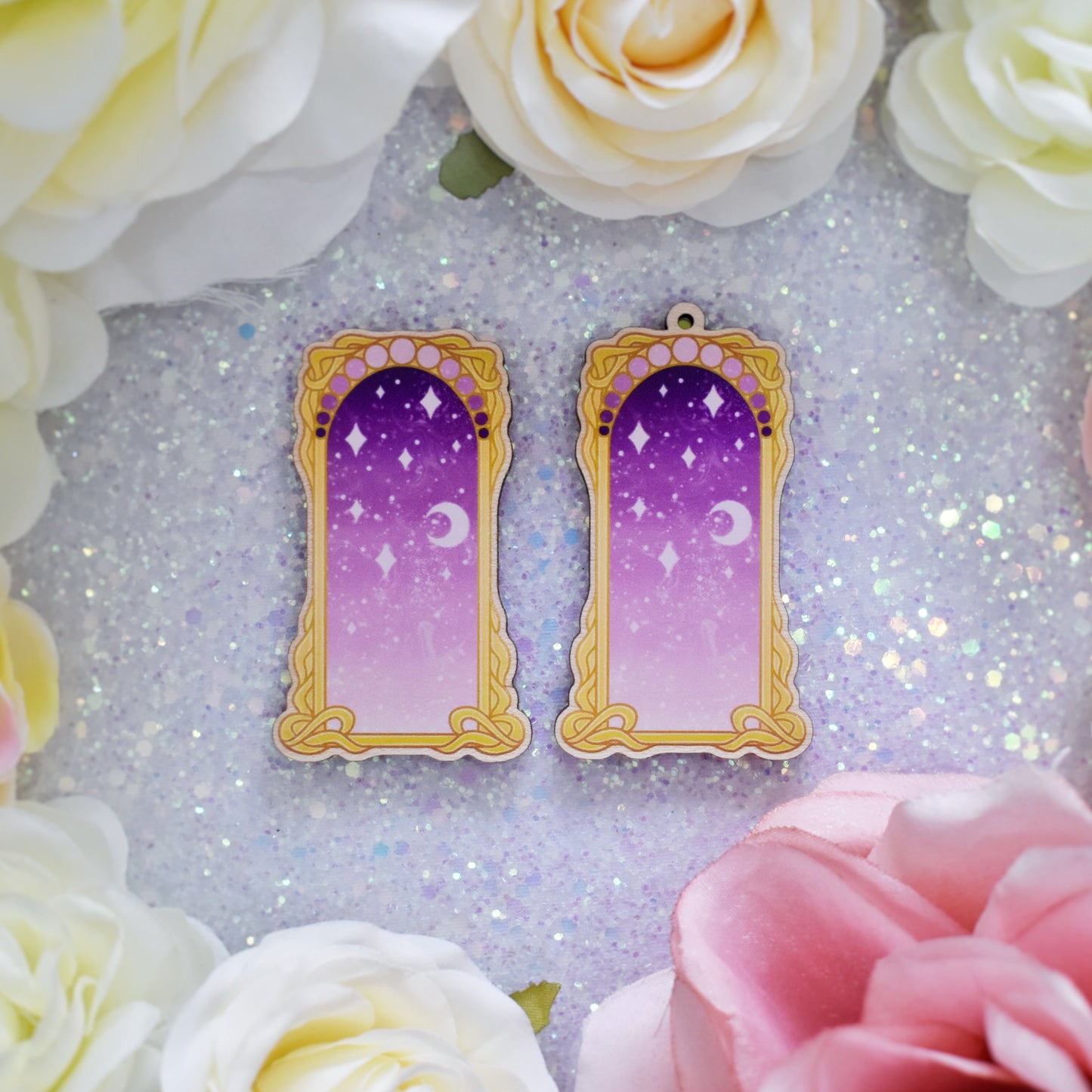 2 wooden charms, 1 with a loop at the top, 1 without next to each other on a sparkly background. Designs have yellow/gold detailed frame. Gradient colour light pink through to darker purpleish pink with stars and a moon inside frame. Fake flowers in white, yellow and pink are visible around edge of photo.