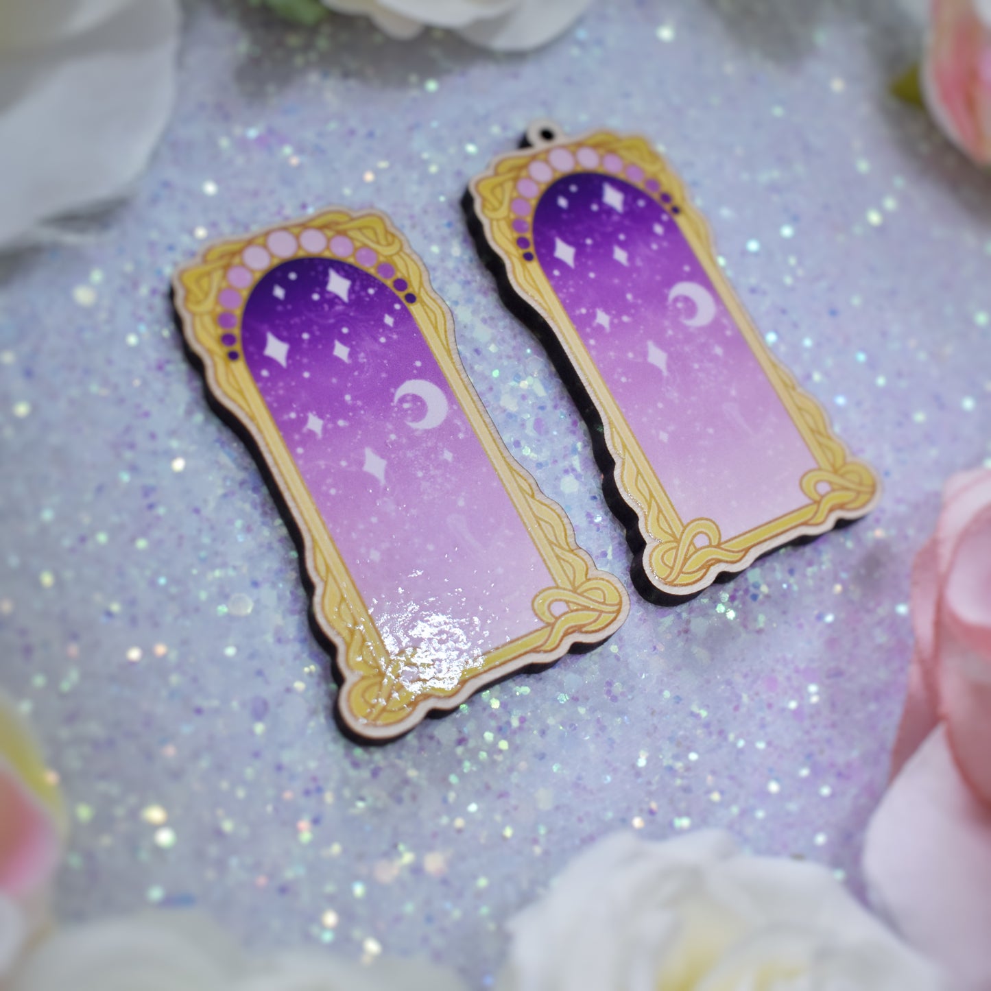 2 wooden charms, 1 with a loop at the top, 1 without next to each other at an angle on a sparkly background. Designs have yellow/gold detailed frame. Gradient colour light pink through to darker purpleish pink with stars and a moon inside frame. Fake flowers in white, yellow and pink are visible around edge of photo.