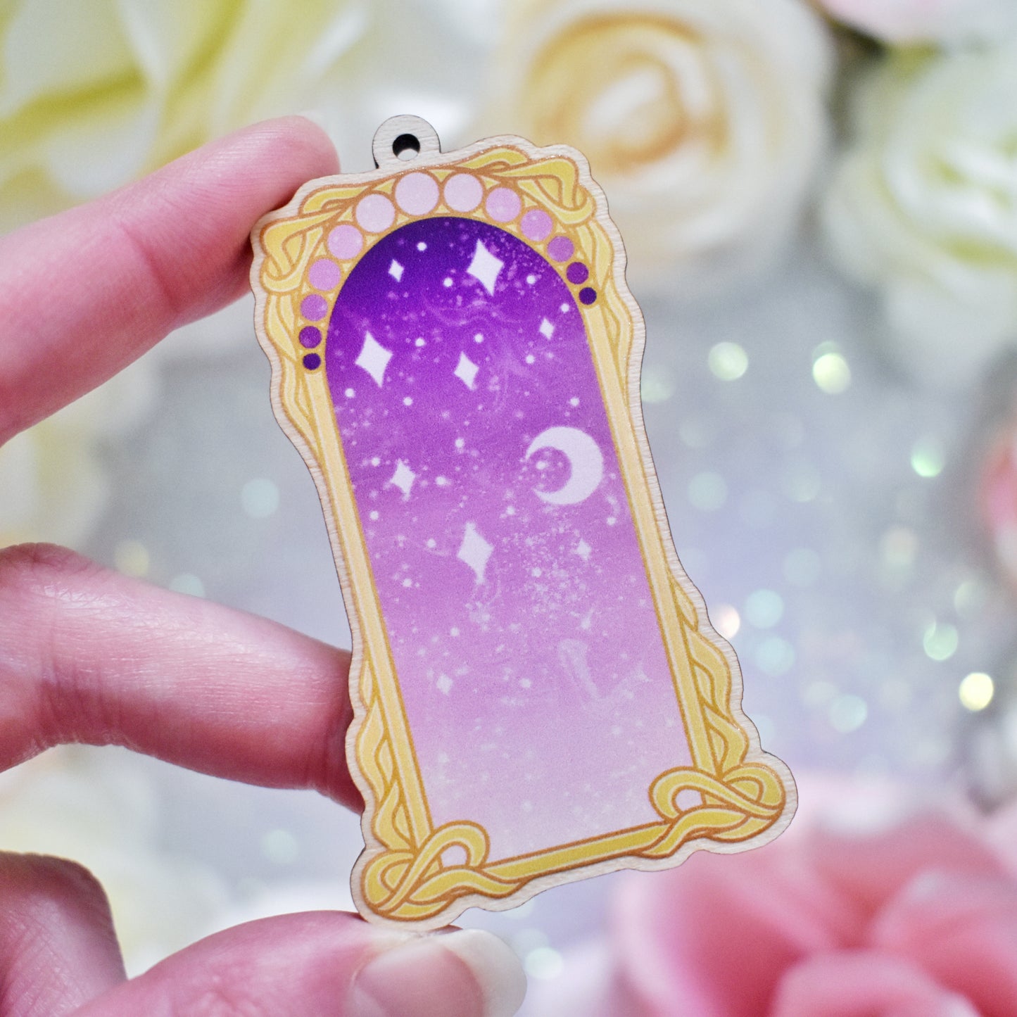 White hand holding wooden charm with top loop. Design has a yellow/gold detailed frame. Gradient colour light pink through to darker purpleish pink with stars and a moon inside frame. Fake flowers in white, yellow and pink are visible around edge of photo.