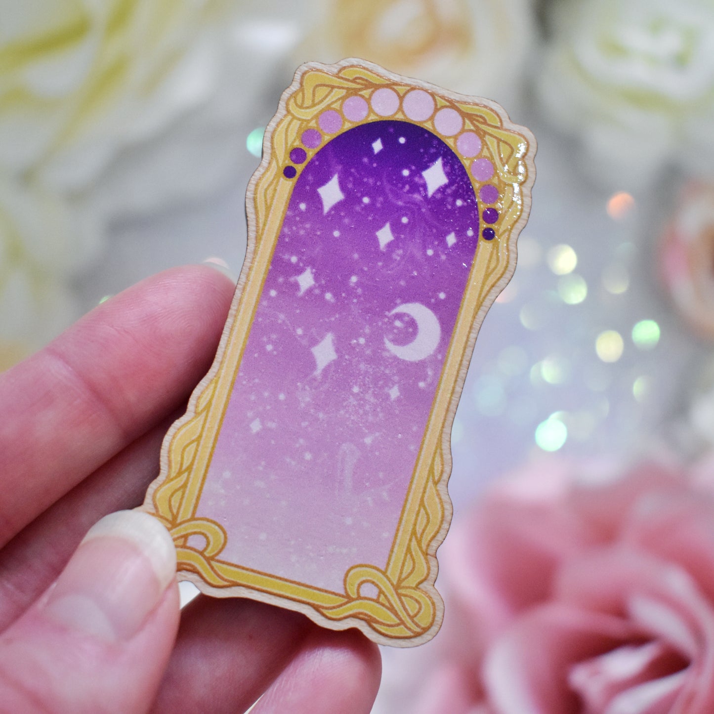 White hand holding wooden charm without top loop. Design has a yellow/gold detailed frame. Gradient colour light pink through to darker purpleish pink with stars and a moon inside frame. Fake flowers in white, yellow and pink are visible around edge of photo.