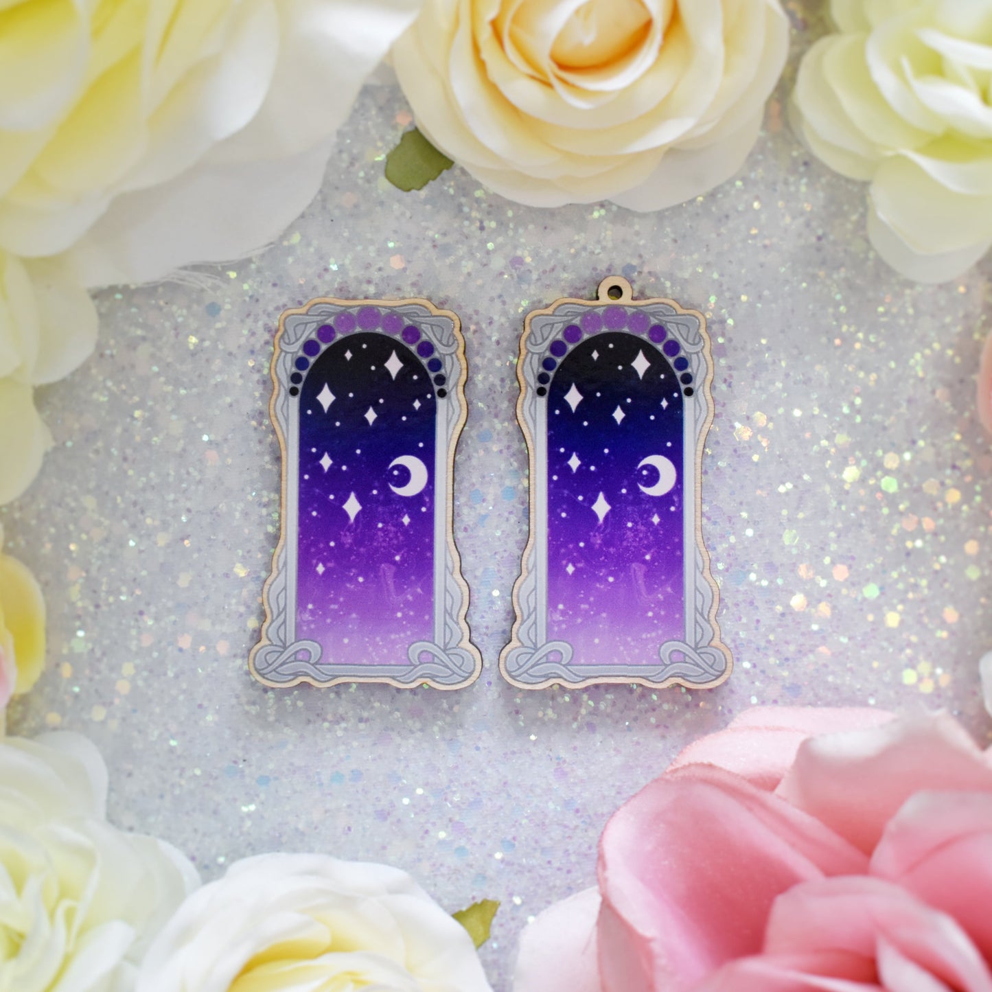 2 wooden charms, 1 with a loop at the top, 1 without next to each other on a sparkly background. Designs have grey detailed frame. Gradient colour yellow pinky purple through to darker bluish purple with stars and a moon inside frame. Fake flowers in white, yellow and pink are visible around edge of photo.
