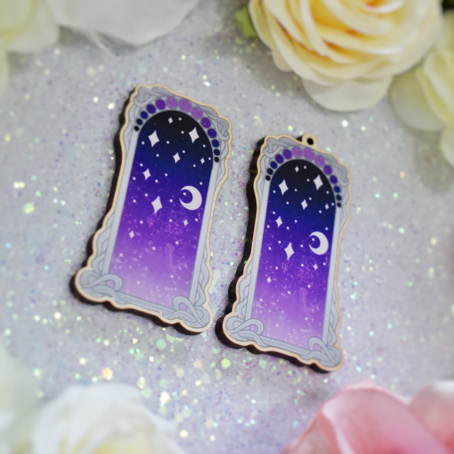2 wooden charms, 1 with a loop at the top, 1 without next to each other at an angle on a sparkly background. Designs have grey detailed frame. Gradient colour yellow pinky purple through to darker bluish purple with stars and a moon inside frame. Fake flowers in white, yellow and pink are visible around edge of photo.