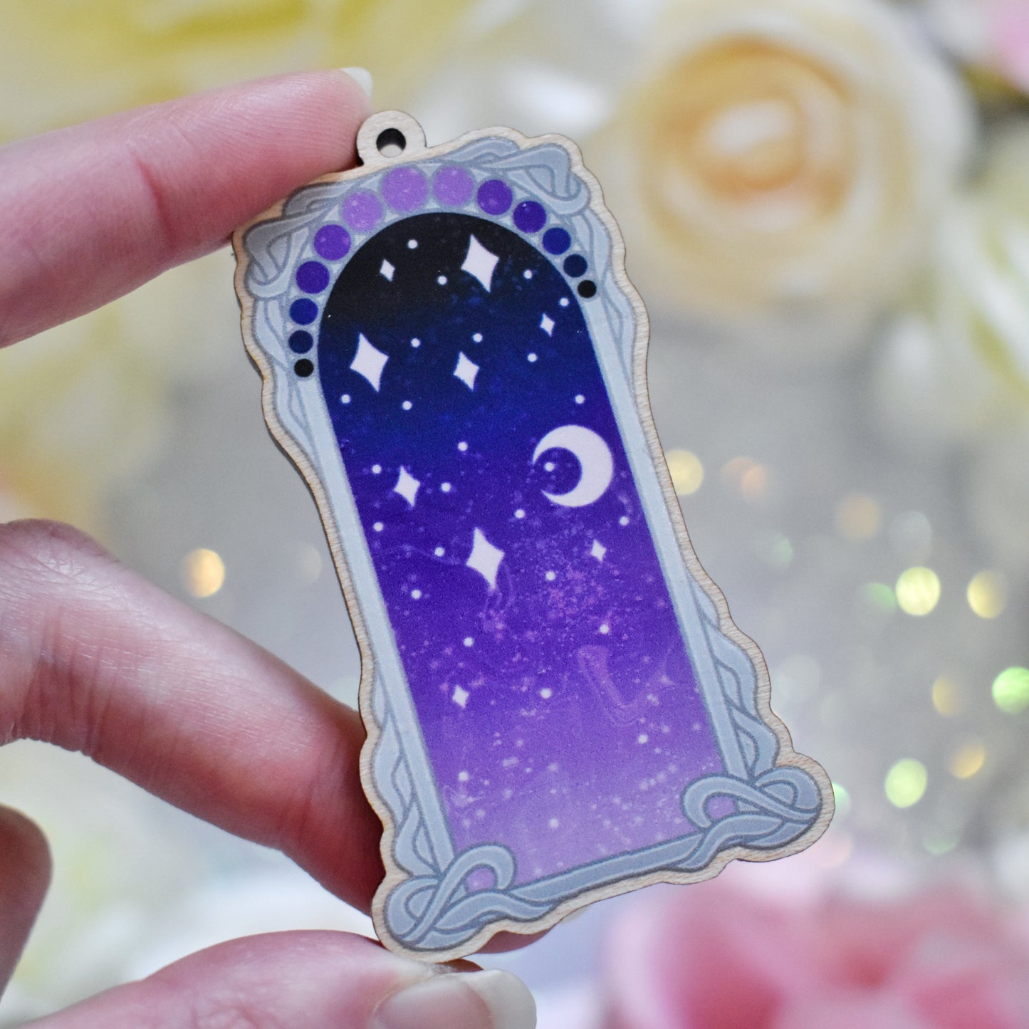 White hand holding wooden charm with top loop. Design has a grey detailed frame. Gradient colour yellow pinky purple through to darker bluish purple with stars and a moon inside frame. Fake flowers in white, yellow and pink are visible around edge of photo.
