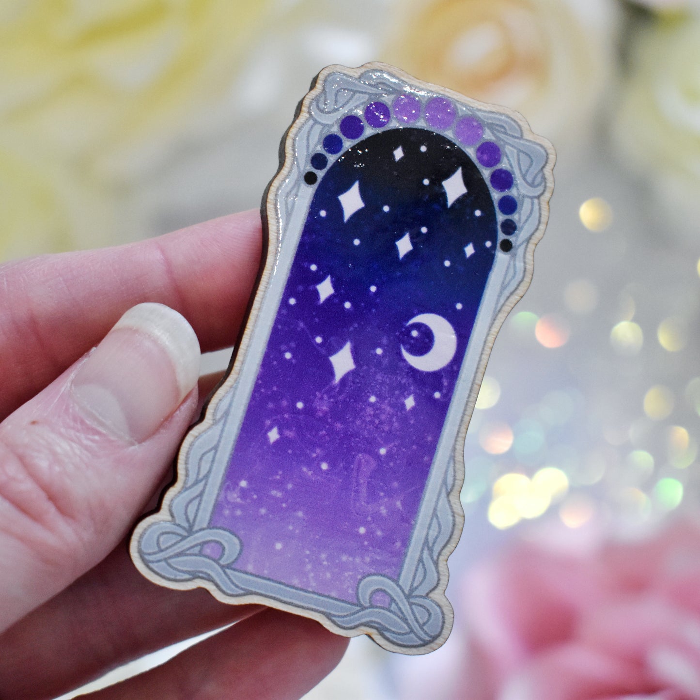 White hand holding wooden charm without top loop. Design has a grey detailed frame. Gradient colour yellow pinky purple through to darker bluish purple with stars and a moon inside frame. Fake flowers in white, yellow and pink are visible around edge of photo.