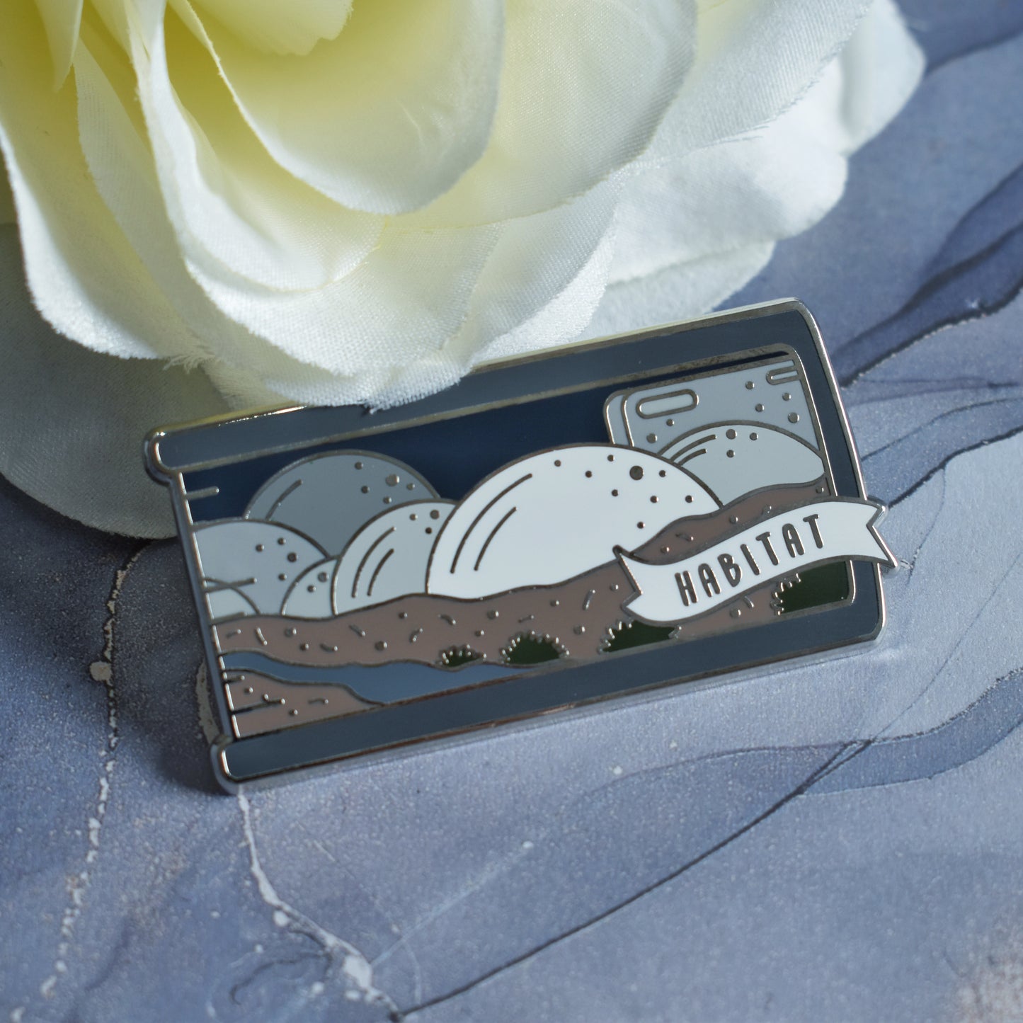 Silver metal enamel pin in centre of image. Enamel pin is shaped like the end of a book in grey and features a scene with different sized half spheres going from white centre front to darker grey the further back they go. Sat on top of brown earth with a river and patches of green plants. A small banner reads habitat on the bottom right of the design. Background of photograph is grey watercolour style with a fake yellow/white flower.