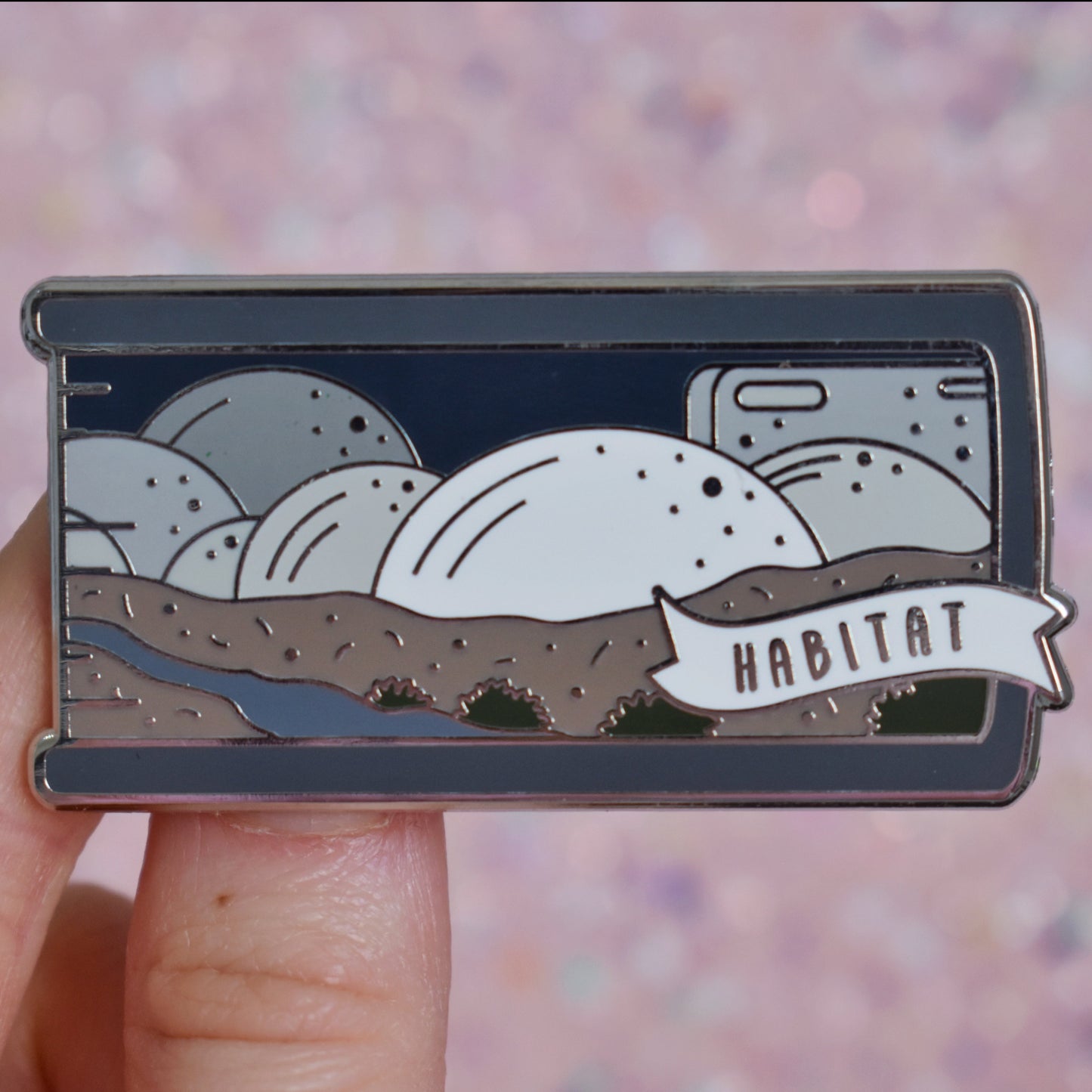 White hand holding silver metal enamel pin. Enamel pin is shaped like the end of a book in grey and features a scene with different sized half spheres going from white centre front to darker grey the further back they go. Sat on top of brown earth with a river and patches of green plants. A small banner reads habitat on the bottom right of the design. Background of photograph is peachy glittery coloured.