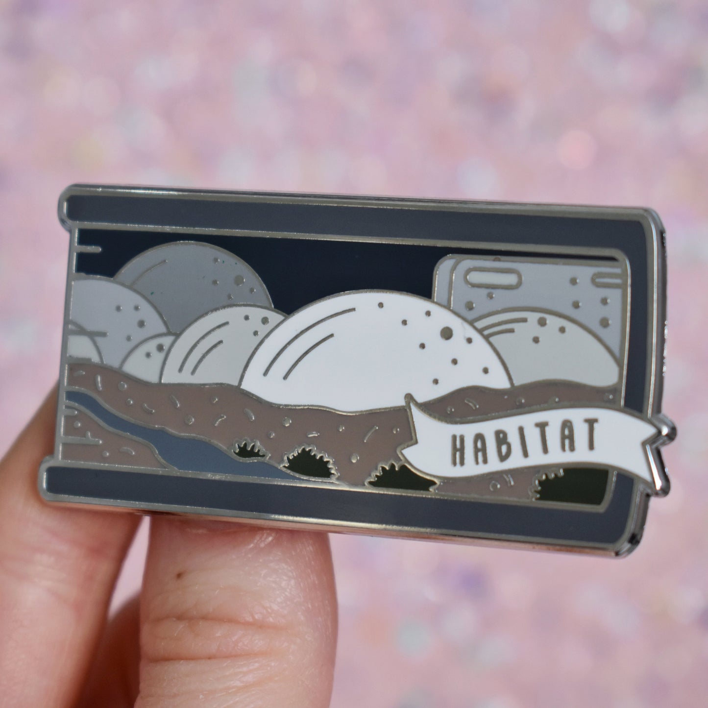 White hand holding enamel pin, tilted to make the silver metal catch more light. Enamel pin is shaped like the end of a book in grey and features a scene with different sized half spheres going from white centre front to darker grey the further back they go. Sat on top of brown earth with a river and patches of green plants. A small banner reads habitat on the bottom right of the design. Background of photograph is peachy glittery coloured.