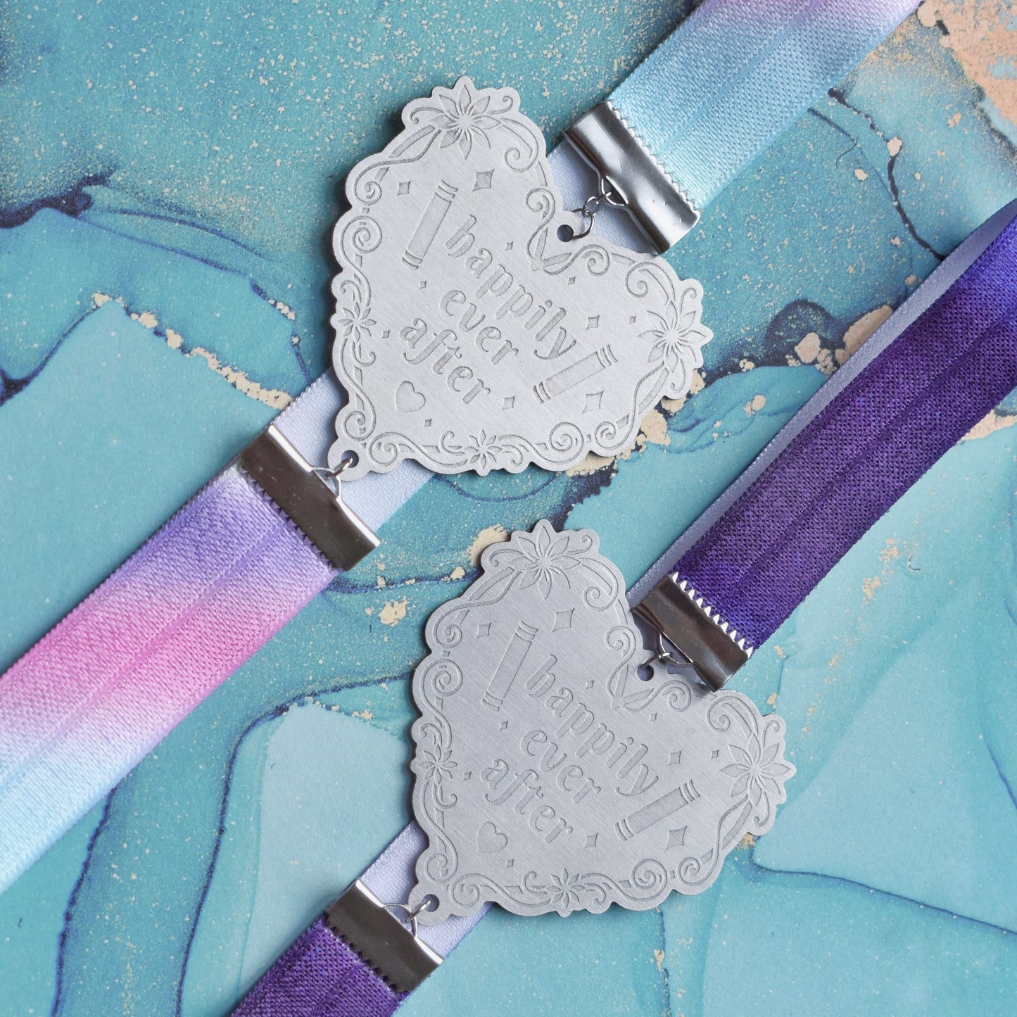 2 book bands at diagonal in the centre of image. Top bookband is pastel rainbow elastic with stainless steel engraved heart and book charm that reads happily ever after. Bottom bookband is purple galaxy elastic with stainless steel engraved heart and book charm that reads happily ever after. On a pale blue/teal watercolour bacground.
